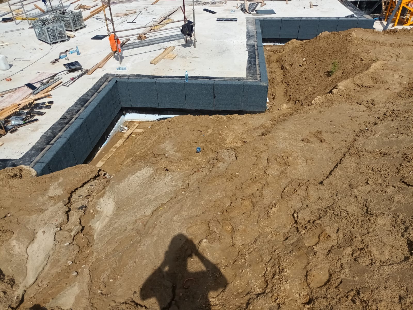 Fundamenty budynku w trakcie izolacji termicznej, widoczne szare płyty izolacyjne. Obok wykop z ziemią i cień fotografa. Na górze rusztowania i materiały budowlane.