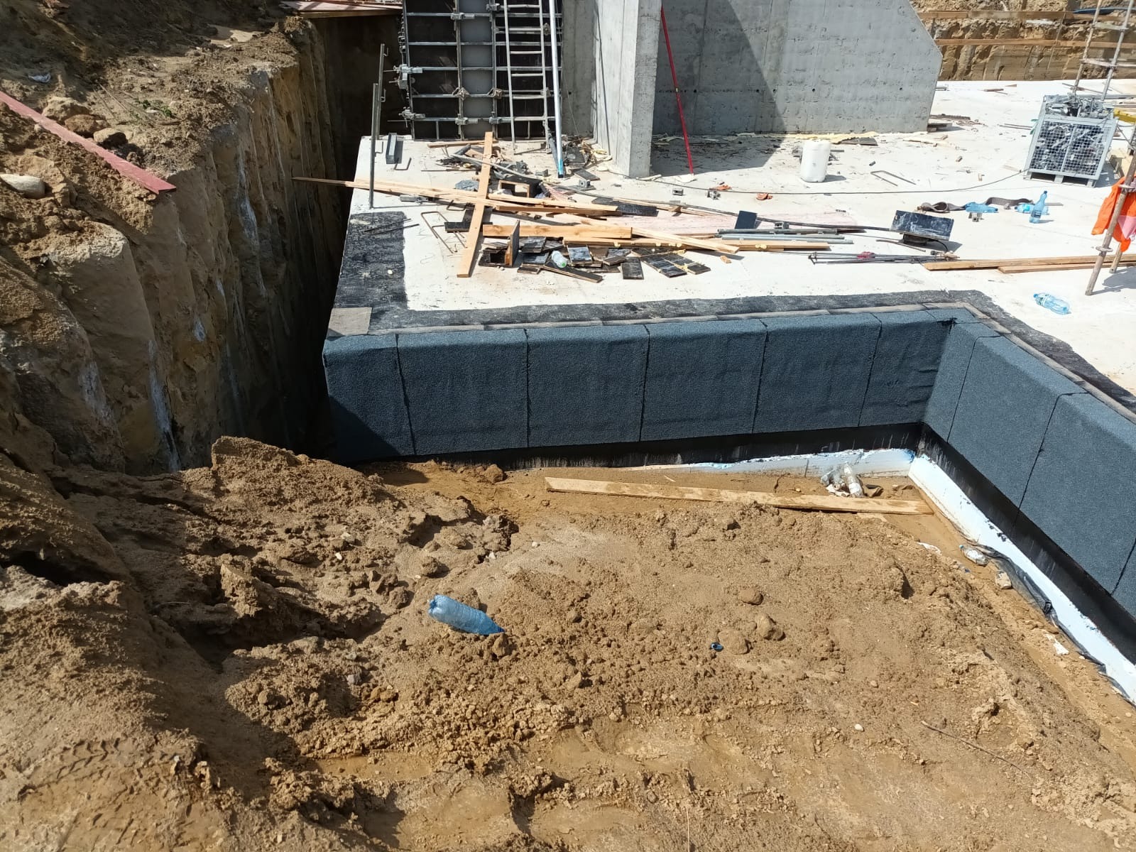 Hydroizolacja fundamentów zabezpieczona płytami izolacyjnymi w wykopie. Widoczne narzędzia i materiały budowlane. Ujęcie z góry ukazuje etap prac.
