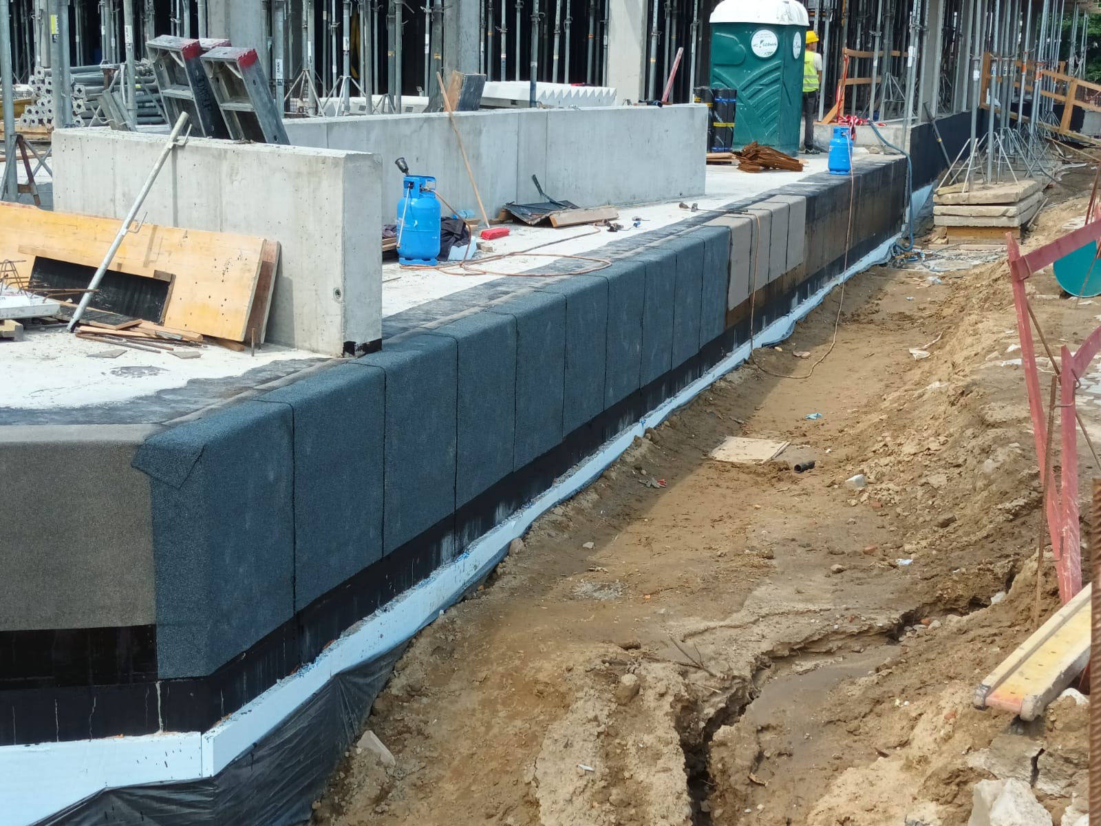 Izolacja przeciwwilgociowa fundamentów budynku w trakcie budowy. Widoczne warstwy izolacyjne i grunt. W tle rusztowania i przenośna toaleta.