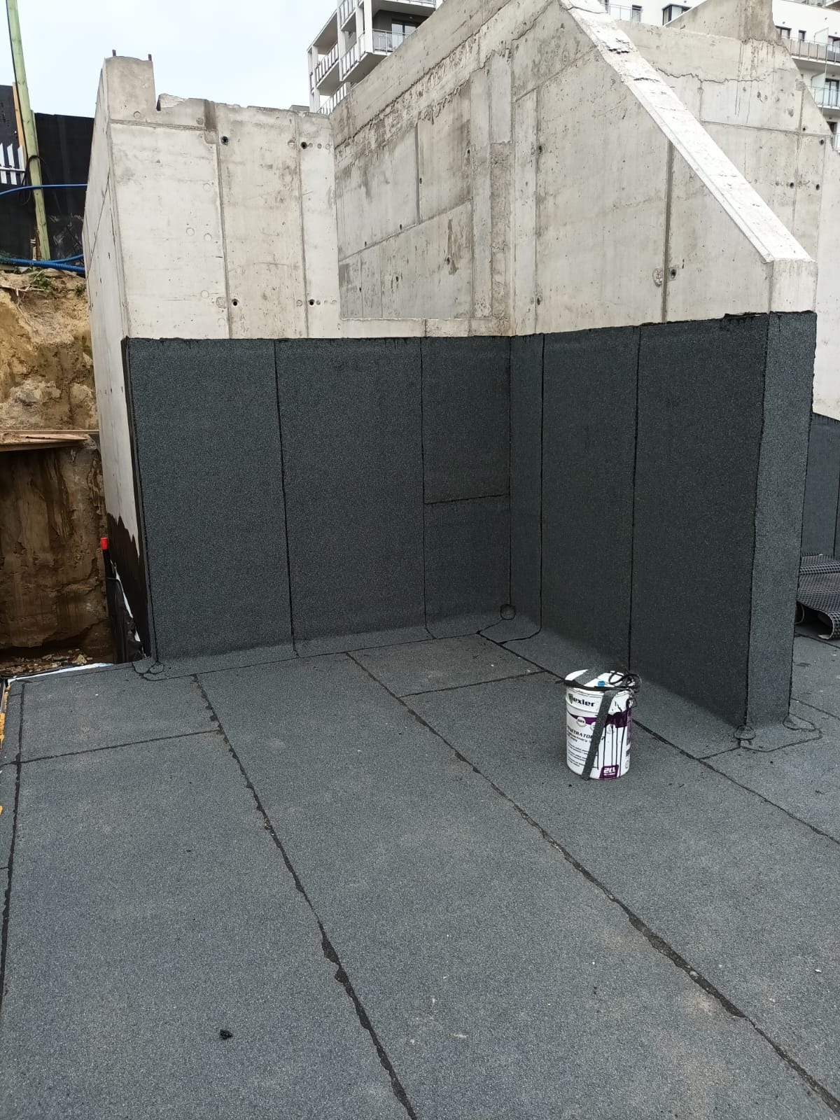 Hydroizolacja fundamentów budynku w trakcie realizacji. Widoczne warstwy izolacyjne na ścianach i podłożu, oraz wiadro z preparatem do hydroizolacji.