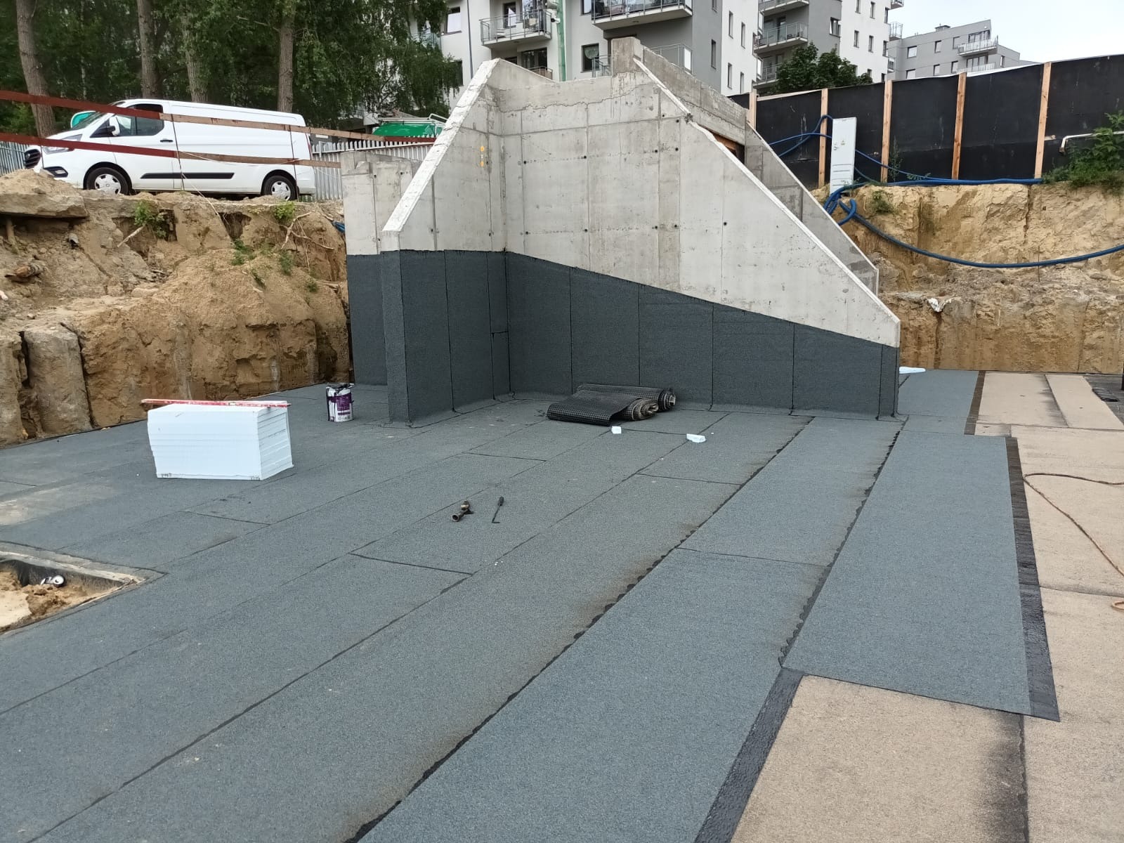 Izolacja fundamentów budynku w trakcie realizacji. Widoczne warstwy izolacyjne na ścianach i podłożu, materiały budowlane i narzędzia. Prace dekarskie.