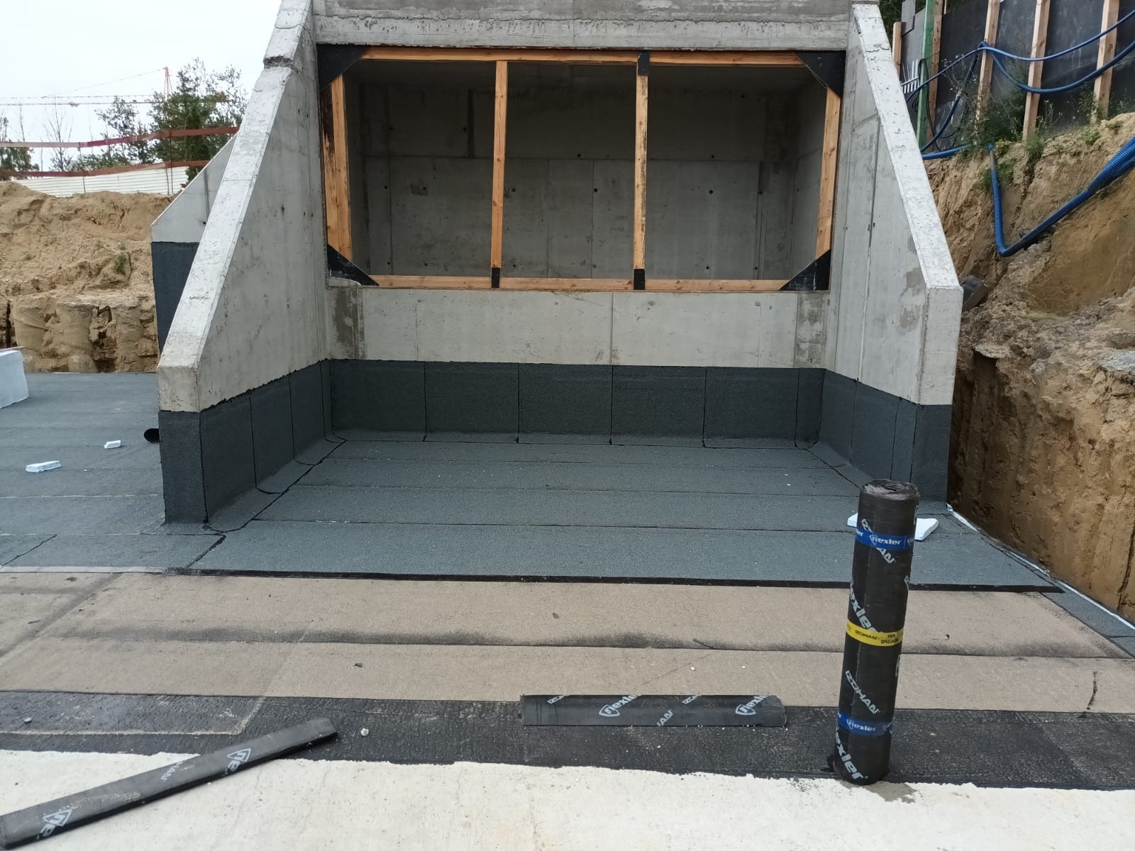 Hydroizolacja fundamentów budynku z betonu komórkowego, z widoczną papą bitumiczną i narzędziami. Wykop ziemny w tle.