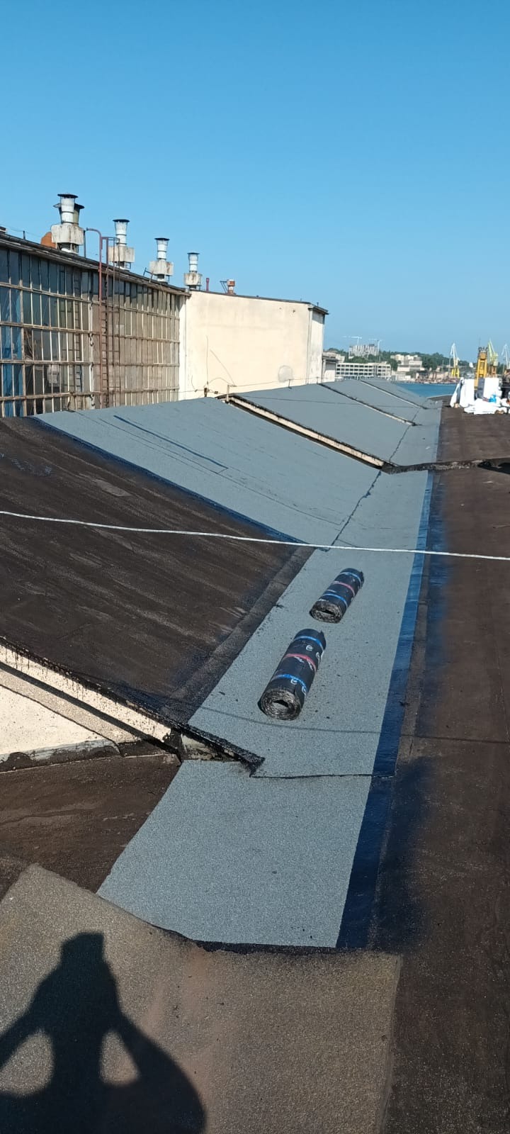 Fragment płaskiego dachu pokrytego nową papą termozgrzewalną w kolorze szarym, z widocznymi rolkami papy. W tle budynki przemysłowe i błękitne niebo.
