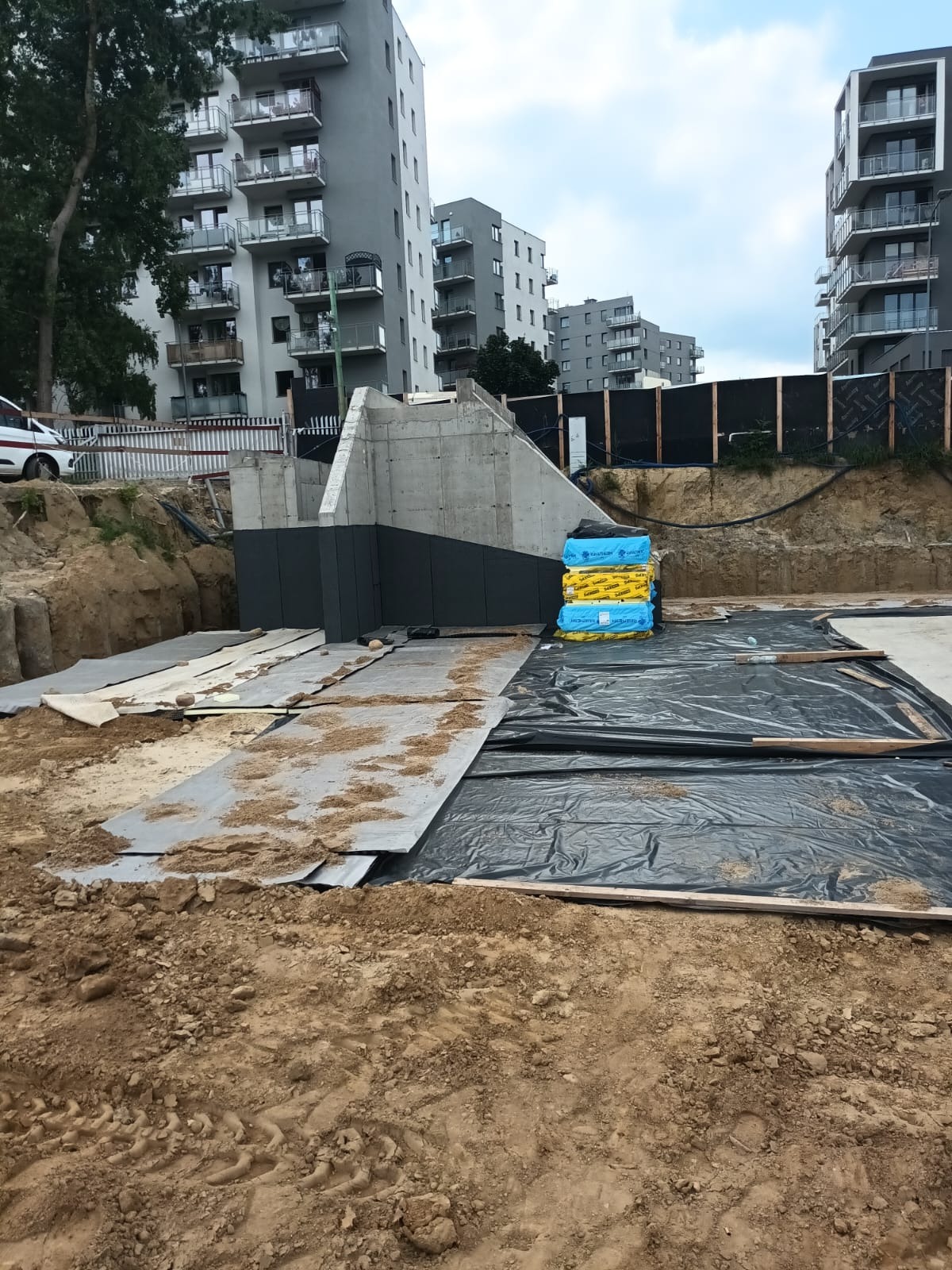 Fundamenty budynku wielorodzinnego z izolacją termiczną, zabezpieczone folią i geowłókniną. W tle bloki mieszkalne.