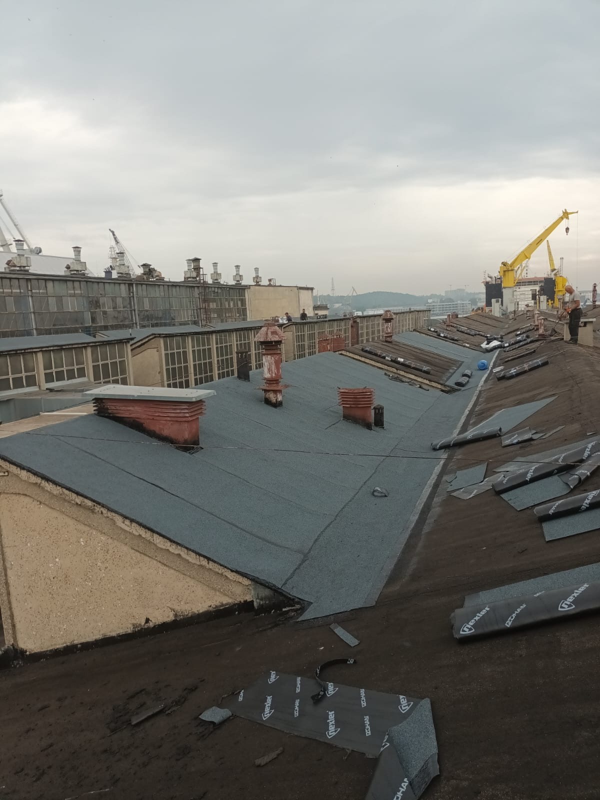 Wykończony fragment dachu z papy termozgrzewalnej w Lubichowie, z widocznymi kominami i częściowo rozłożonymi rolkami papy. W tle budynki przemysłowe i dźwigi.