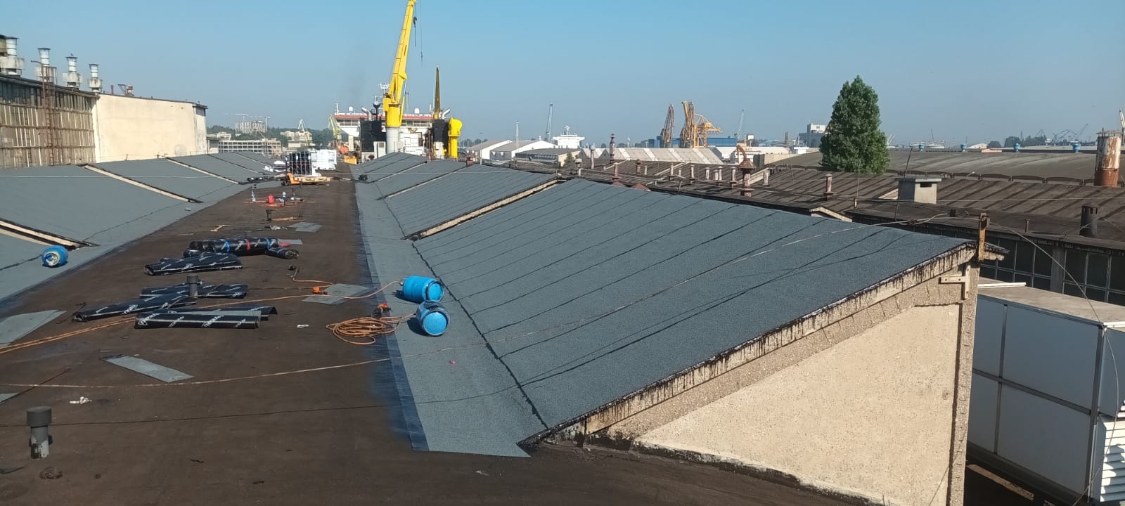Prace dekarskie na rozległym dachu przemysłowym. Widoczne rolki papy, butle gazowe i częściowo pokryta powierzchnia. W tle dźwigi portowe i zabudowa miejska.