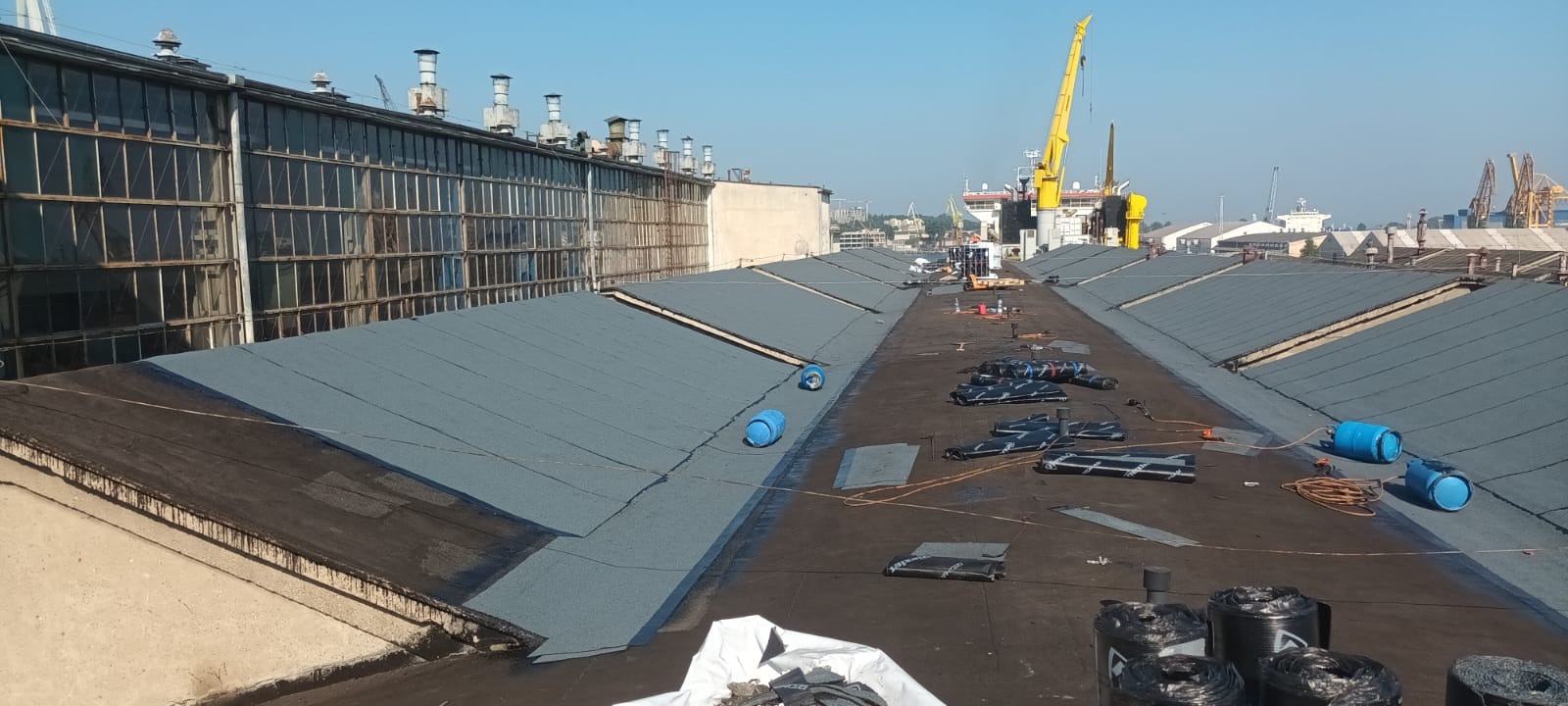 Prace na dachu hali przemysłowej: częściowo położona papa termozgrzewalna, butle z gazem, widok na port z dźwigami w tle. Dach płaski z widocznymi kominami wentylacyjnymi.