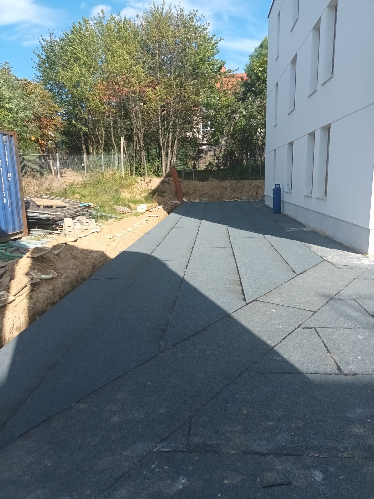 Ułożona, szara nawierzchnia z płyt gumowych na zewnątrz budynku, obok wykopu. W tle drzewa i fragment białej ściany. Widoczny fragment niebieskiego kontenera.