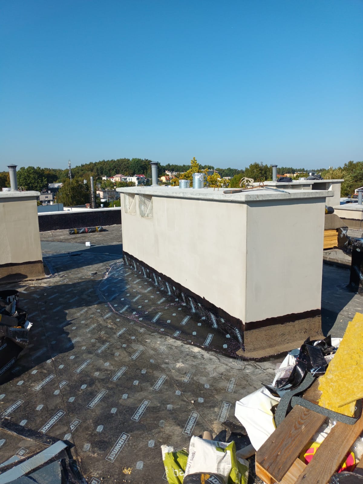 Prace na dachu płaskim: izolacja bitumiczna wokół kominów wentylacyjnych. Widoczne rolki papy i narzędzia. Błękitne niebo w tle.
