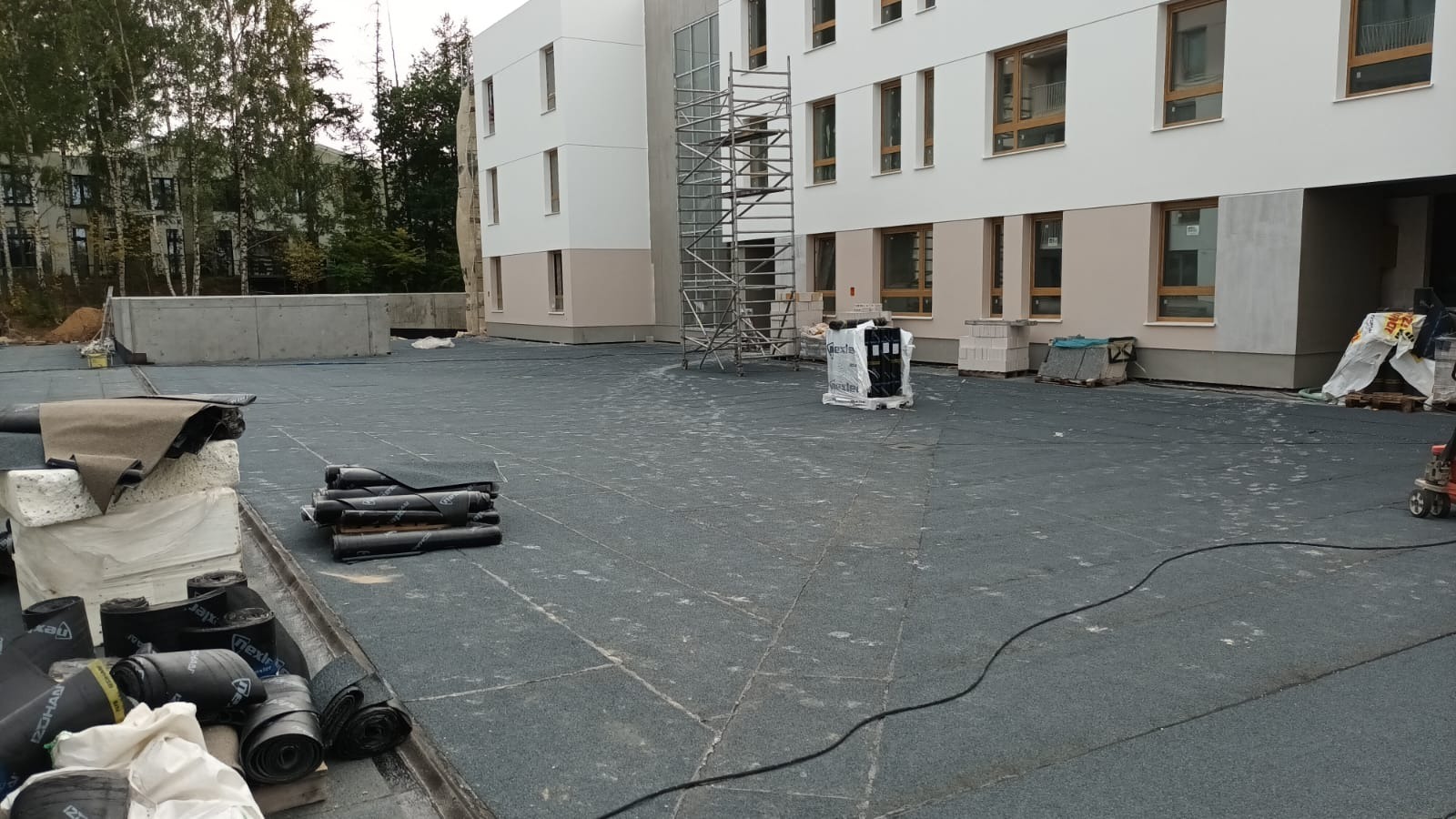 Wykończony, płaski dach budynku pokryty papą termozgrzewalną. Zrolowane arkusze papy leżą na powierzchni. W tle rusztowanie i elewacja budynku.