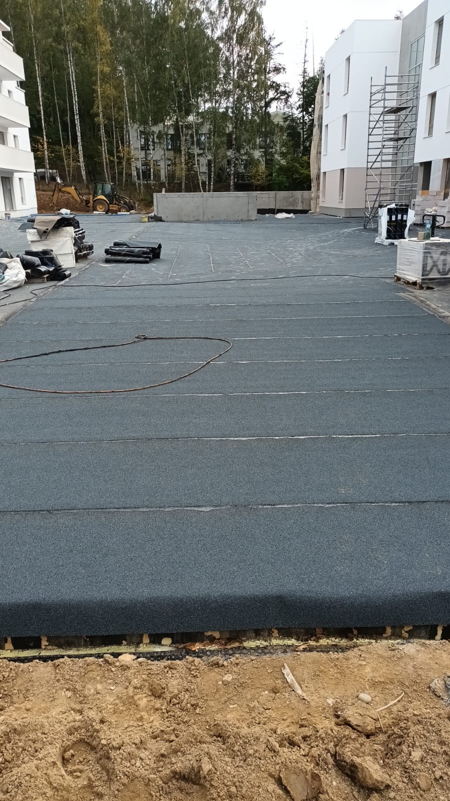 Położona papa na płaskim dachu budynku wielorodzinnego w trakcie budowy. Widoczne materiały budowlane, ziemia i koparka w tle.