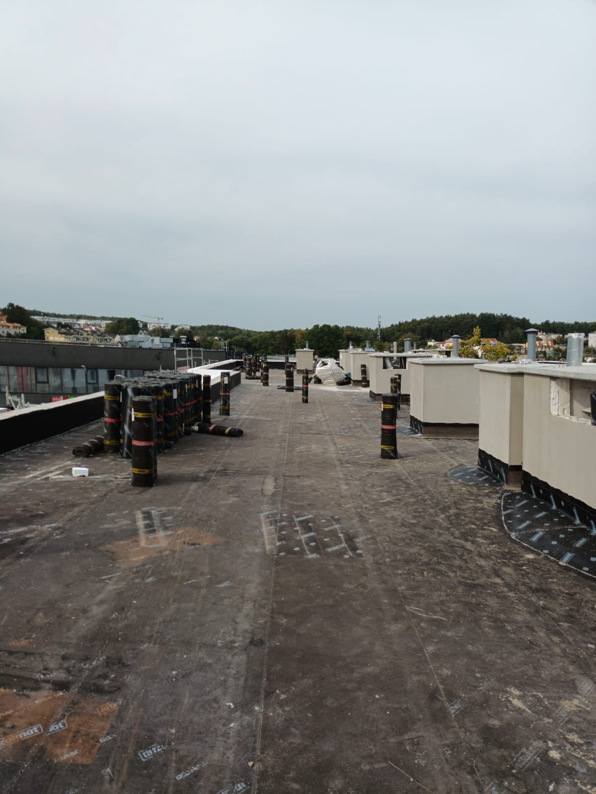 Widok na płaski dach z rolkami papy i elementami wentylacyjnymi. Prace dekarskie w toku, widoczne materiały izolacyjne i kominy wentylacyjne. Tło stanowi zabudowa miejska.
