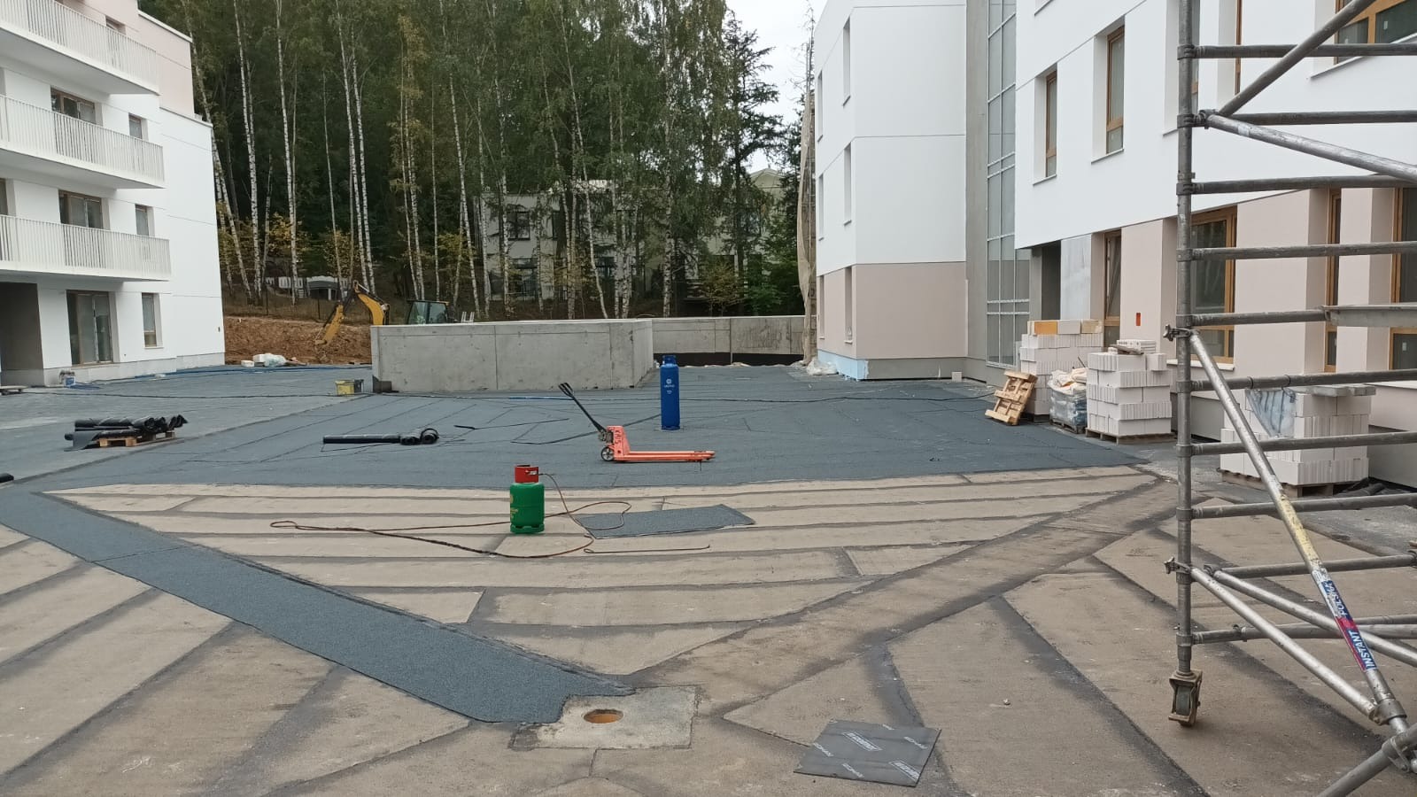 Prace na dachu płaskim budynku wielorodzinnego: rozłożona papa, butle gazowe, paleta z bloczkami i rusztowanie. W tle drzewa i budynki.