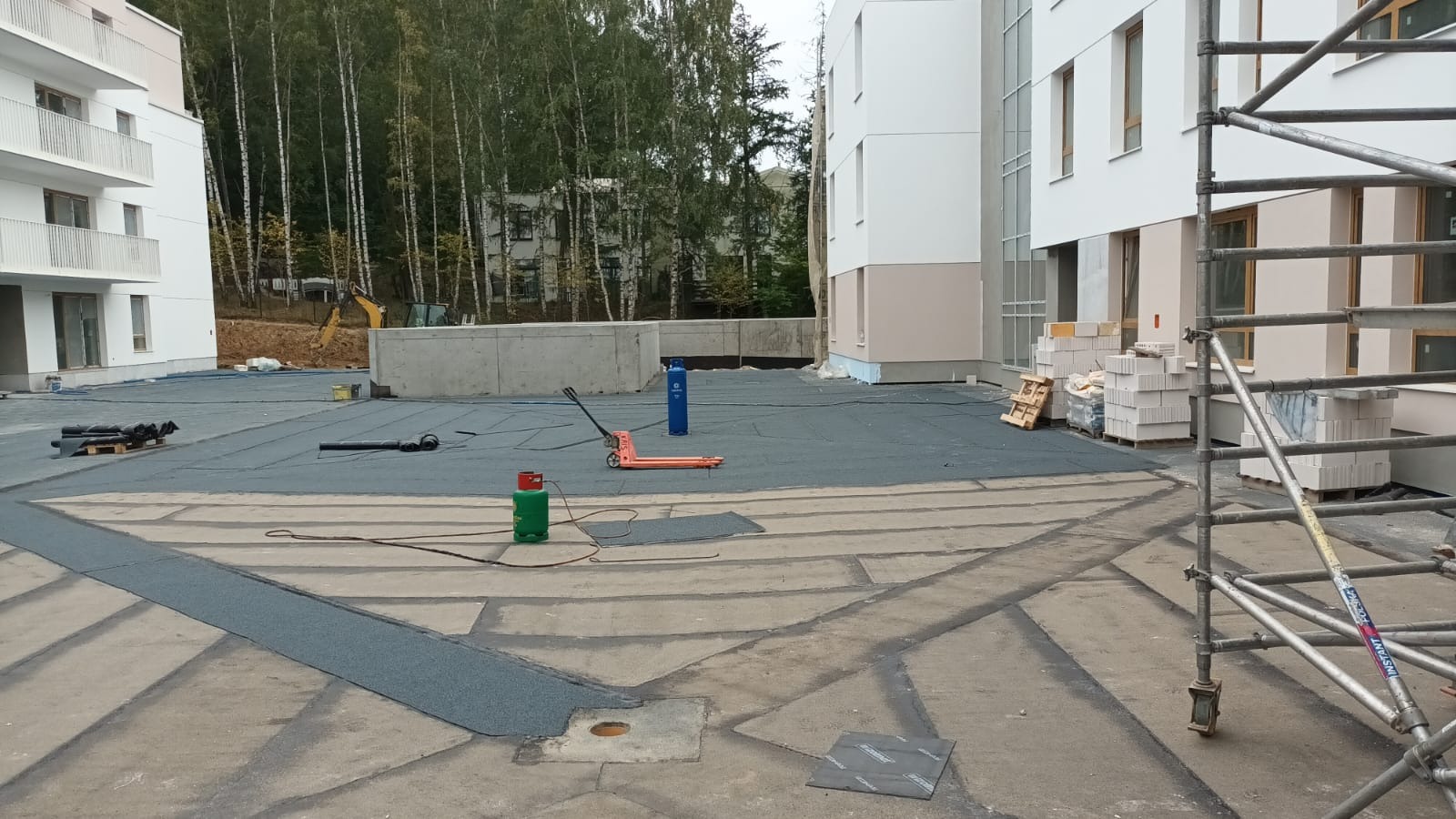 Etap prac na dachu budynku wielorodzinnego: hydroizolacja papą termozgrzewalną, materiały budowlane, rusztowanie, butle z gazem i wózek paletowy na dachu. Widok z góry.