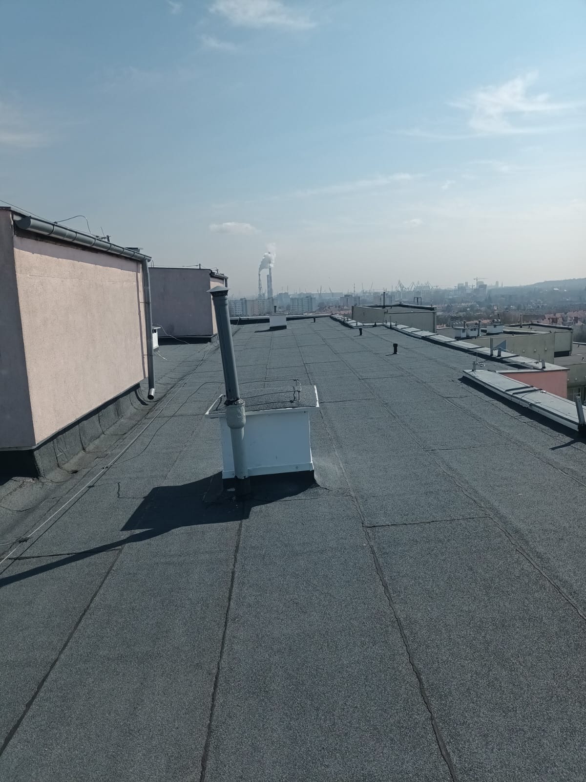 Wykończony dach pokryty papą na budynku z widocznymi kominami wentylacyjnymi i zabudową. W tle panorama miasta i kominy przemysłowe. Słoneczny dzień.
