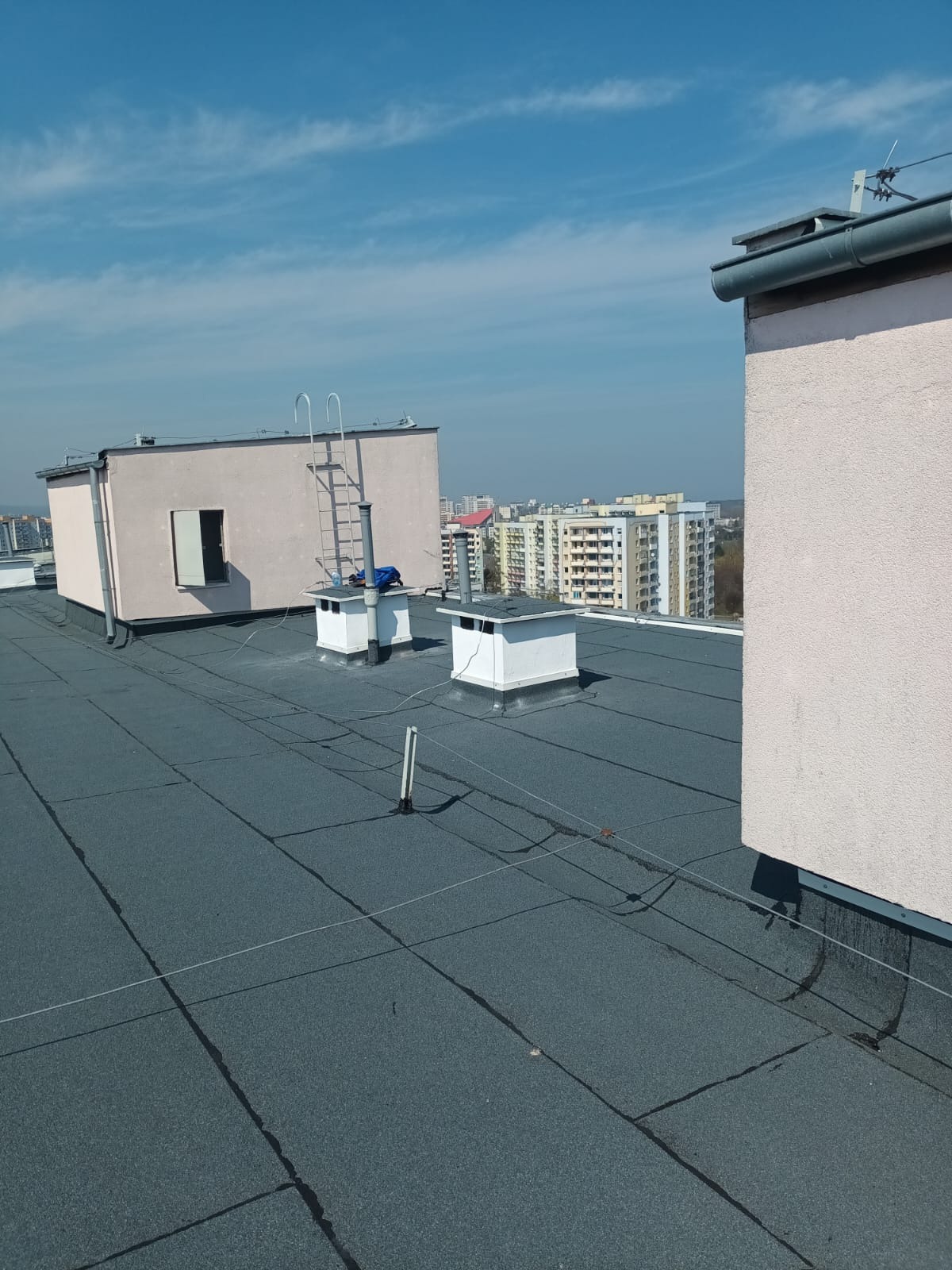 Szary dach pokryty papą termozgrzewalną z kominami wentylacyjnymi i drabiną w słoneczny dzień. W tle zabudowa miejska.