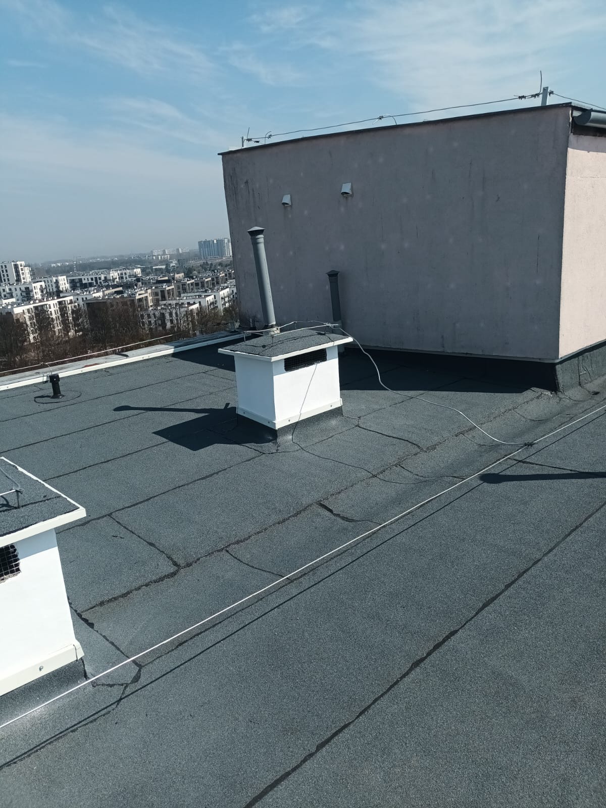 Szary dach pokryty papą z kominkami wentylacyjnymi na tle zabudowy miejskiej w słoneczny dzień. Widoczne pęknięcia na papie.