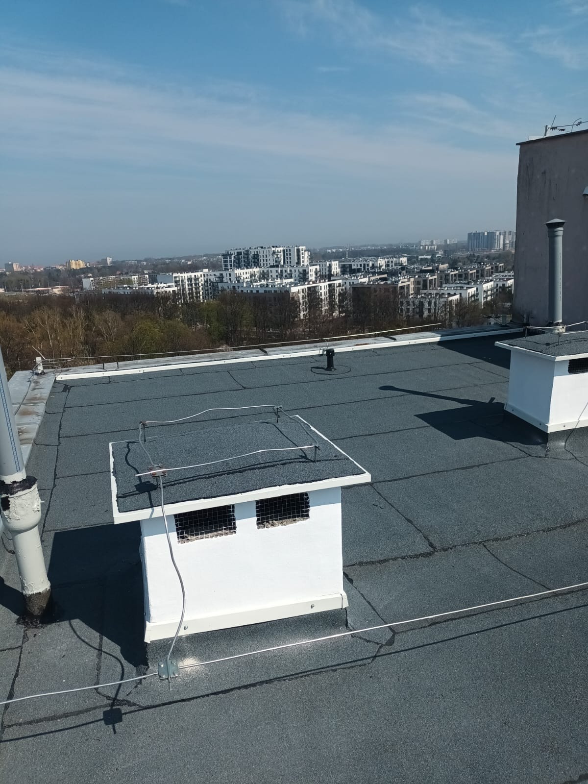 Płaski dach pokryty papą z elementami wentylacyjnymi i odgromowymi na tle zabudowy miejskiej w słoneczny dzień. Widok z góry.