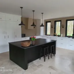 Nowoczesna kuchnia z czarną wyspą, drewnianą misą i kwiatami. Białe szafki, okna z czarnymi ramami i trawertynowa podłoga tworzą eleganckie wnętrze.