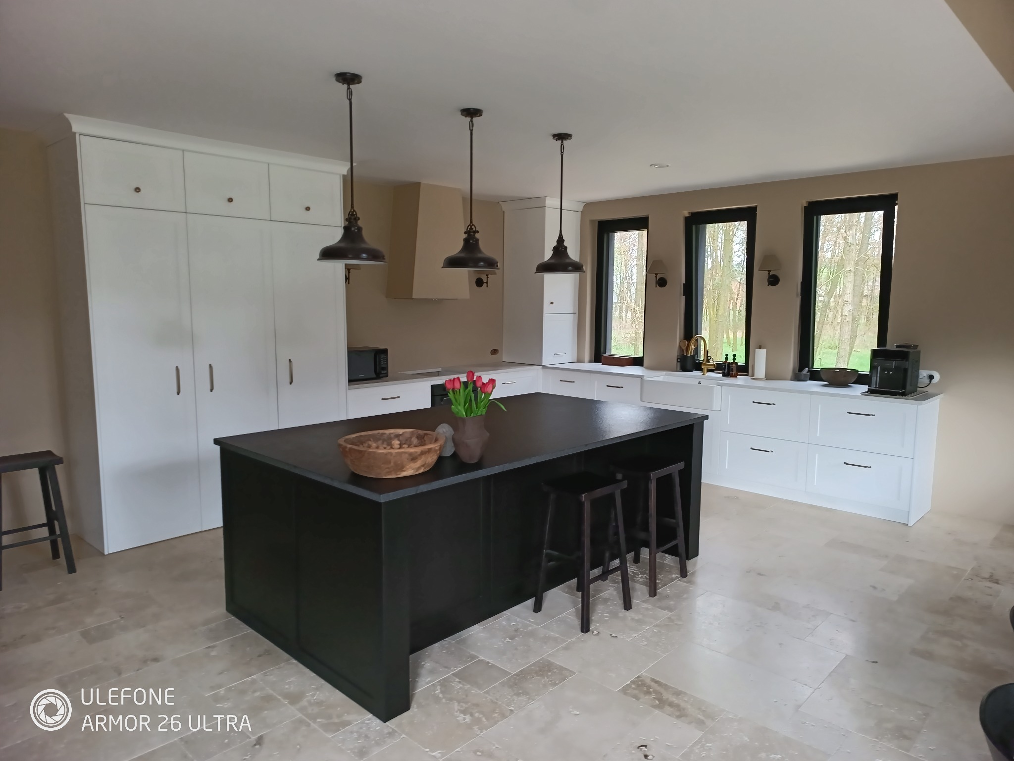 Nowoczesna kuchnia z czarną wyspą, drewnianą misą i kwiatami. Białe szafki, okna z czarnymi ramami i trawertynowa podłoga tworzą eleganckie wnętrze.