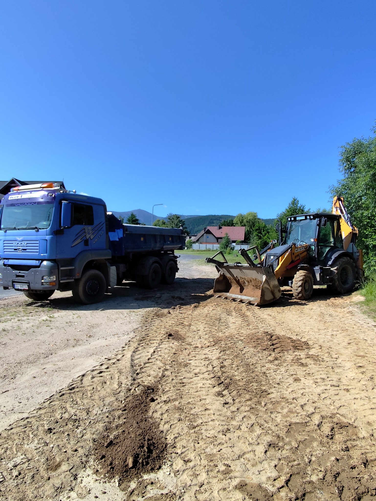 Niebieska ciężarówka MAN obok koparko-ładowarki CASE na piaszczystym terenie. W tle zabudowania i błękitne niebo. Widoczne ślady opon.