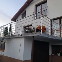 Dom z balkonem i schodami zewnętrznymi, zabezpieczonymi metalową balustradą. Widoczne okna z brązowymi ramami i elementy roślinności. Ujęcie z dołu.