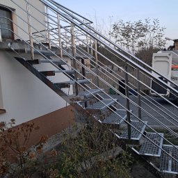 INOX STYLE Paulina Suter-Gładki - Zewnętrzne schody metalowe z ażurowymi stopniami i poręczami ze stali nierdzewnej, zamontowane przy ścianie budynku. W tle widoczny samochód dostawczy.