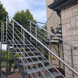 INOX STYLE Paulina Suter-Gładki - Zewnętrzne schody metalowe z połyskującą balustradą, prowadzące do wejścia budynku z elewacją z kamienia. W tle zieleń drzew i krzewów, błękitne niebo.
