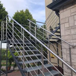 Zewnętrzne schody metalowe z połyskującą balustradą, prowadzące do wejścia budynku z elewacją z kamienia. W tle zieleń drzew i krzewów, błękitne niebo.