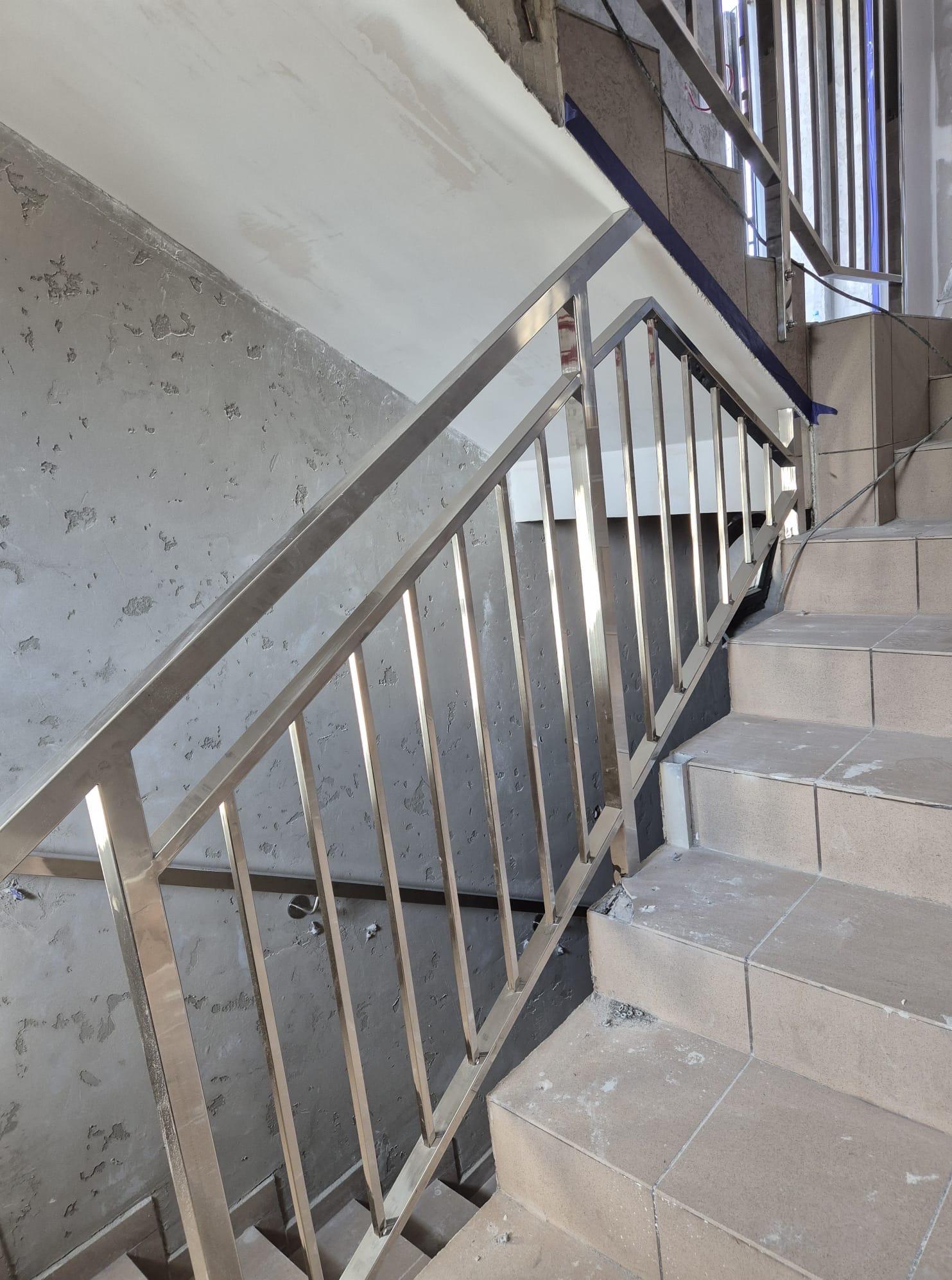 Montaż poręczy ze stali nierdzewnej na schodach w trakcie wykańczania. Widoczne płytki schodowe i surowe ściany. Poręcz o prostym designie, z pionowymi słupkami.