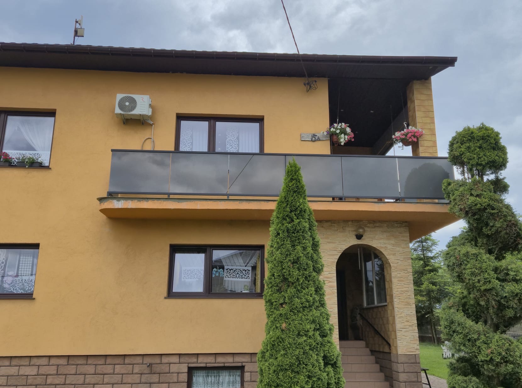 Żółty dom z balkonem z szarą, przeszkloną balustradą, na której wiszą doniczki z kwiatami. Widoczne schody wejściowe z poręczą. Uroku dodają iglaki.