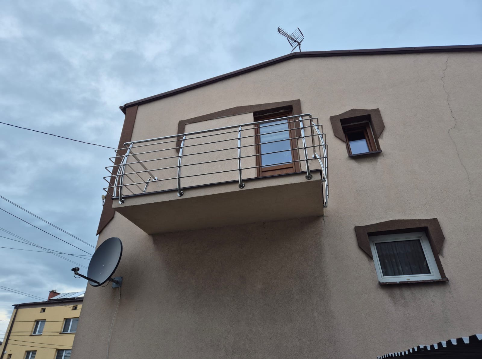 Kanciasty balkon z prostą, metalową balustradą na tle beżowej elewacji budynku. Widoczna antena satelitarna i okna o nietypowych obramowaniach.