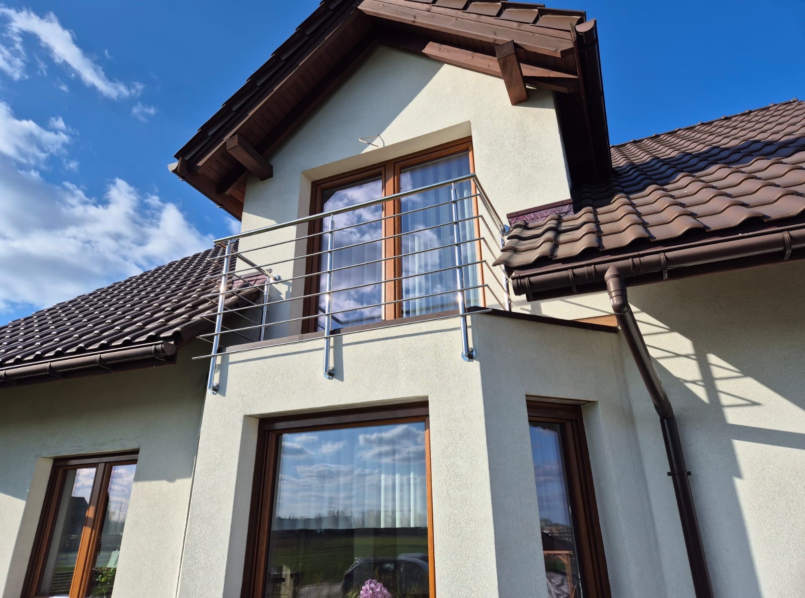 Dom z beżową elewacją i balkonem z prostą, nowoczesną, metalową balustradą. W oknach odbija się błękitne niebo z chmurami. Dach brązowy, dwuspadowy.