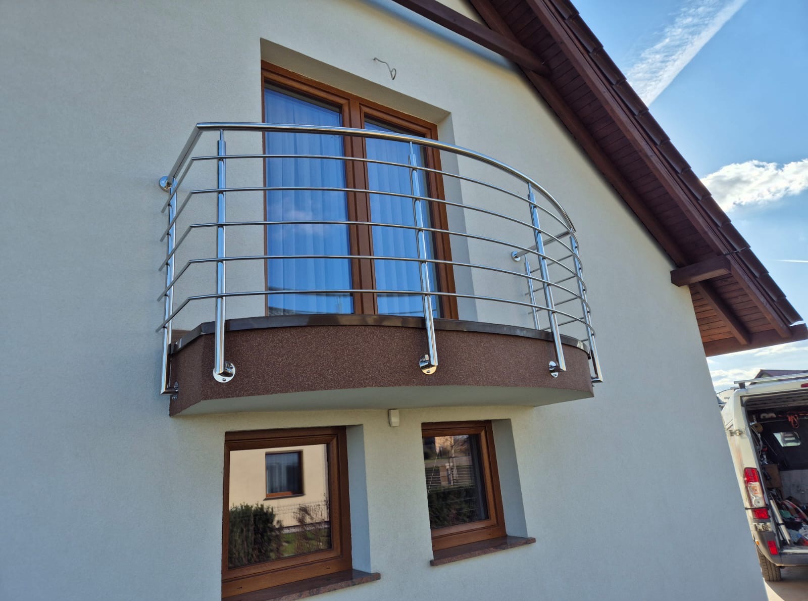 Balkon z zaokrągloną, nowoczesną balustradą ze stali nierdzewnej, zamontowaną na elewacji budynku z drewnianymi oknami w słoneczny dzień.