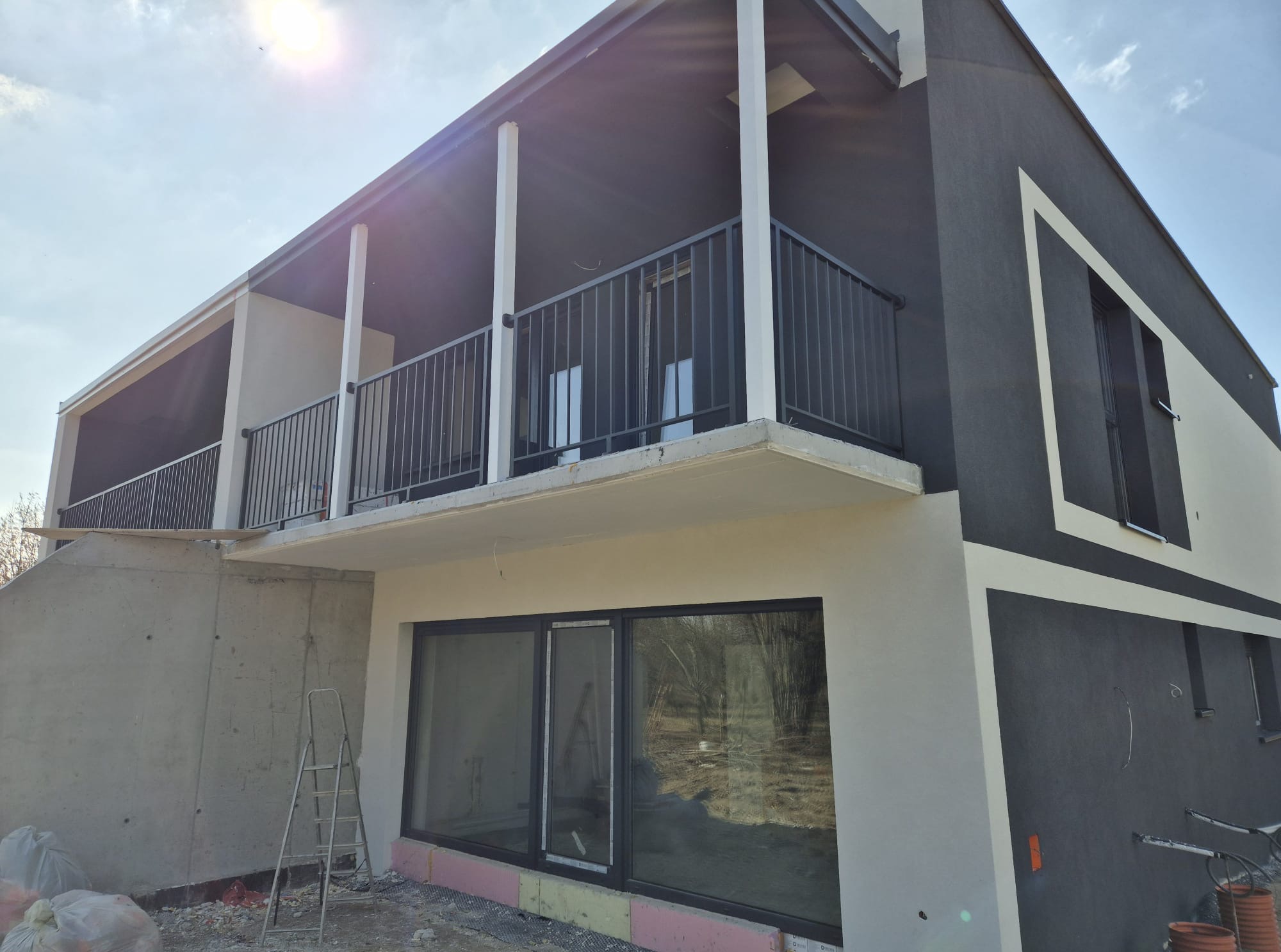 Nowoczesny dom z balkonem zabezpieczonym czarną, metalową balustradą. Widoczne białe kolumny podtrzymujące zadaszenie i surowe wykończenie elewacji.