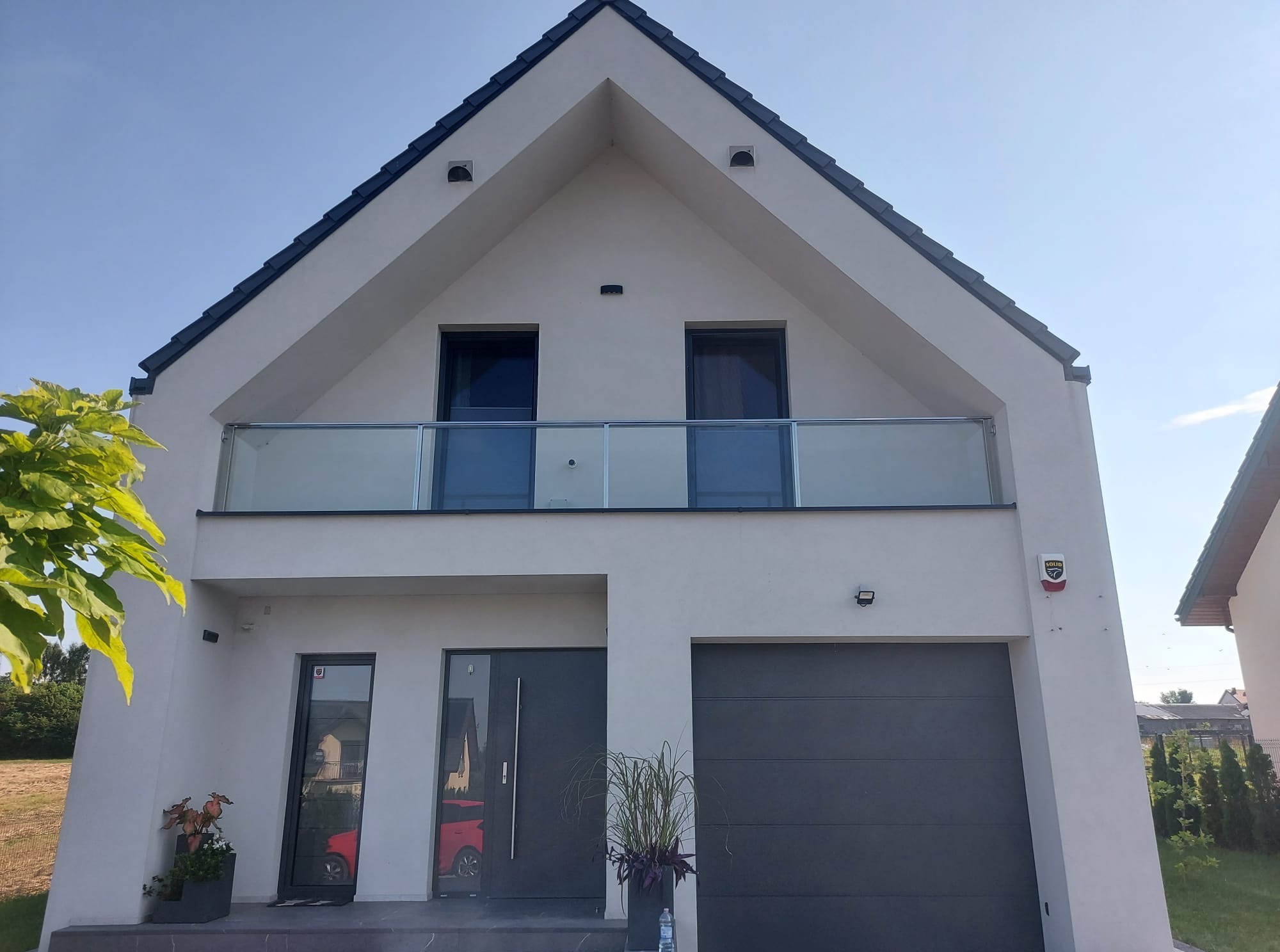 Nowoczesny dom jednorodzinny w Słupi z balkonem zabezpieczonym szkłem, szara stolarka okienna i drzwiowa, garaż z bramą segmentową, minimalistyczny styl.