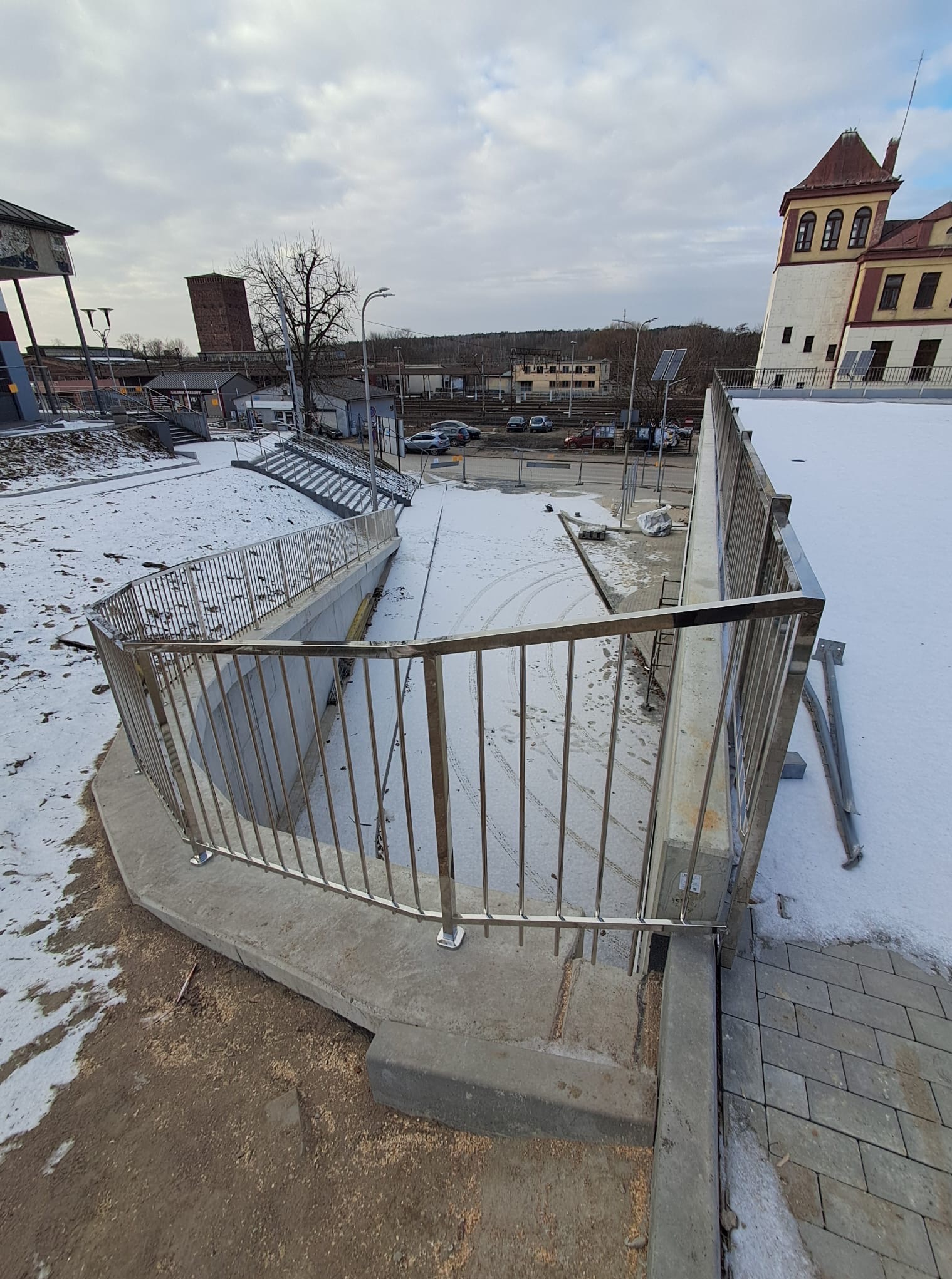 Nierdzewna balustrada schodowa w przestrzeni miejskiej, betonowe schody, fragment placu pokrytego śniegiem, w tle budynki i pochmurne niebo.