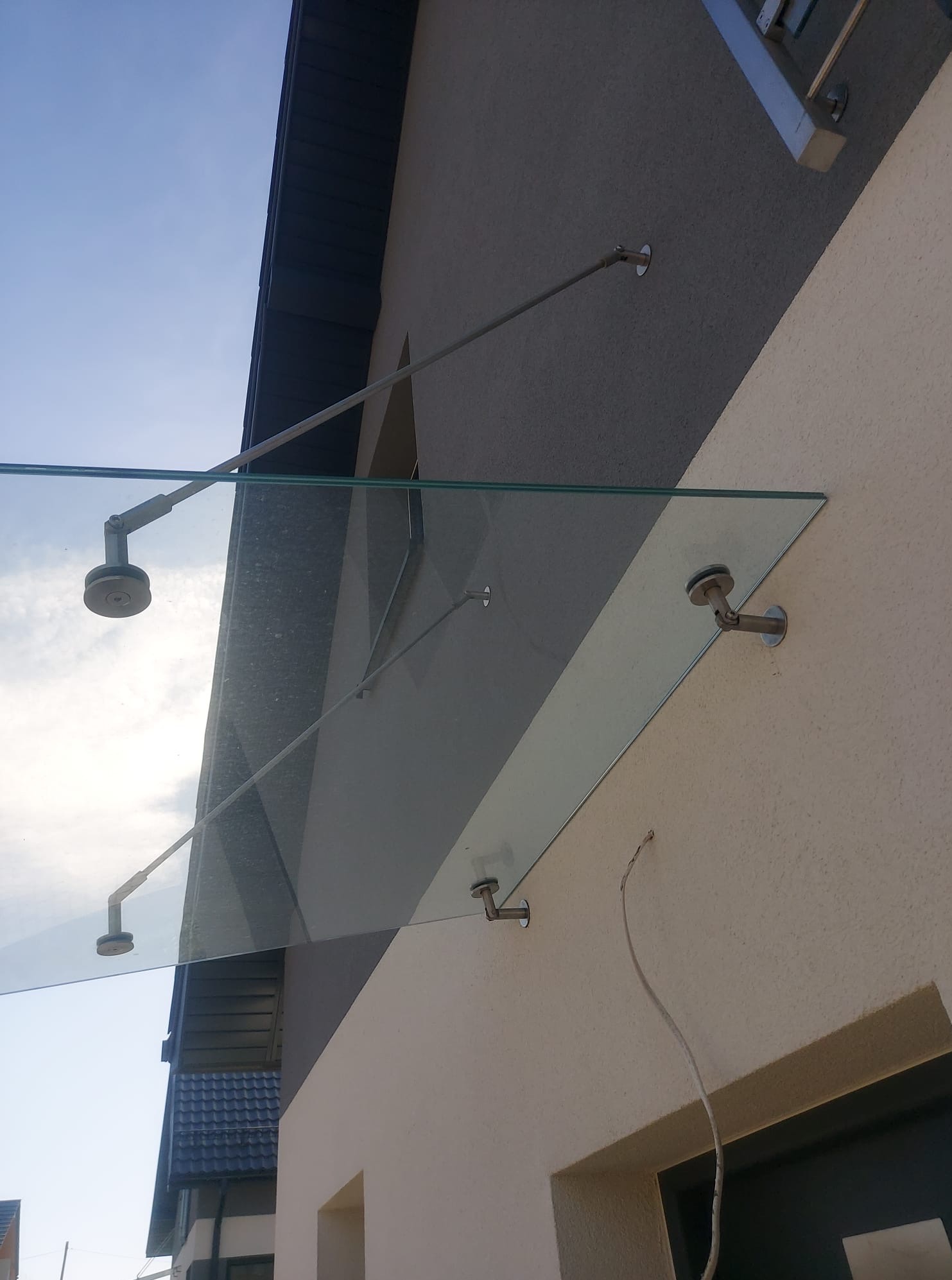 Szklany daszek nad wejściem do budynku, mocowany na metalowych wspornikach. Widok z dołu, niebo w tle. Nowoczesne rozwiązanie architektoniczne.