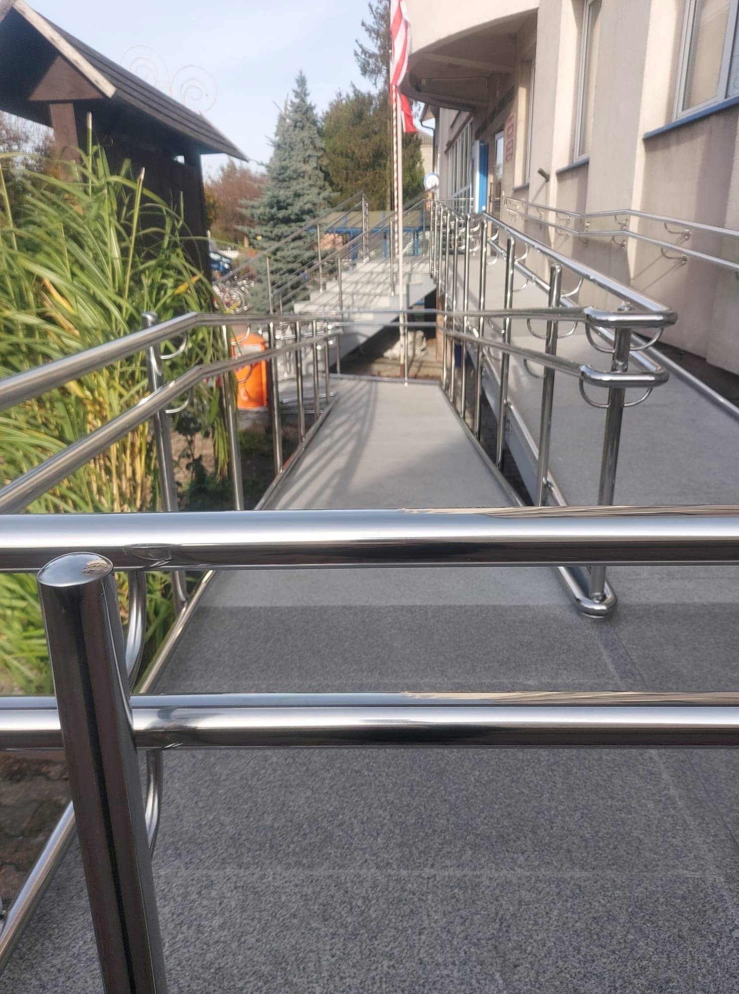 Rampa wejściowa z polerowanymi, metalowymi poręczami, ułatwiająca dostęp do budynku. Widok z dołu, z bliska na detale konstrukcyjne i fakturę nawierzchni.