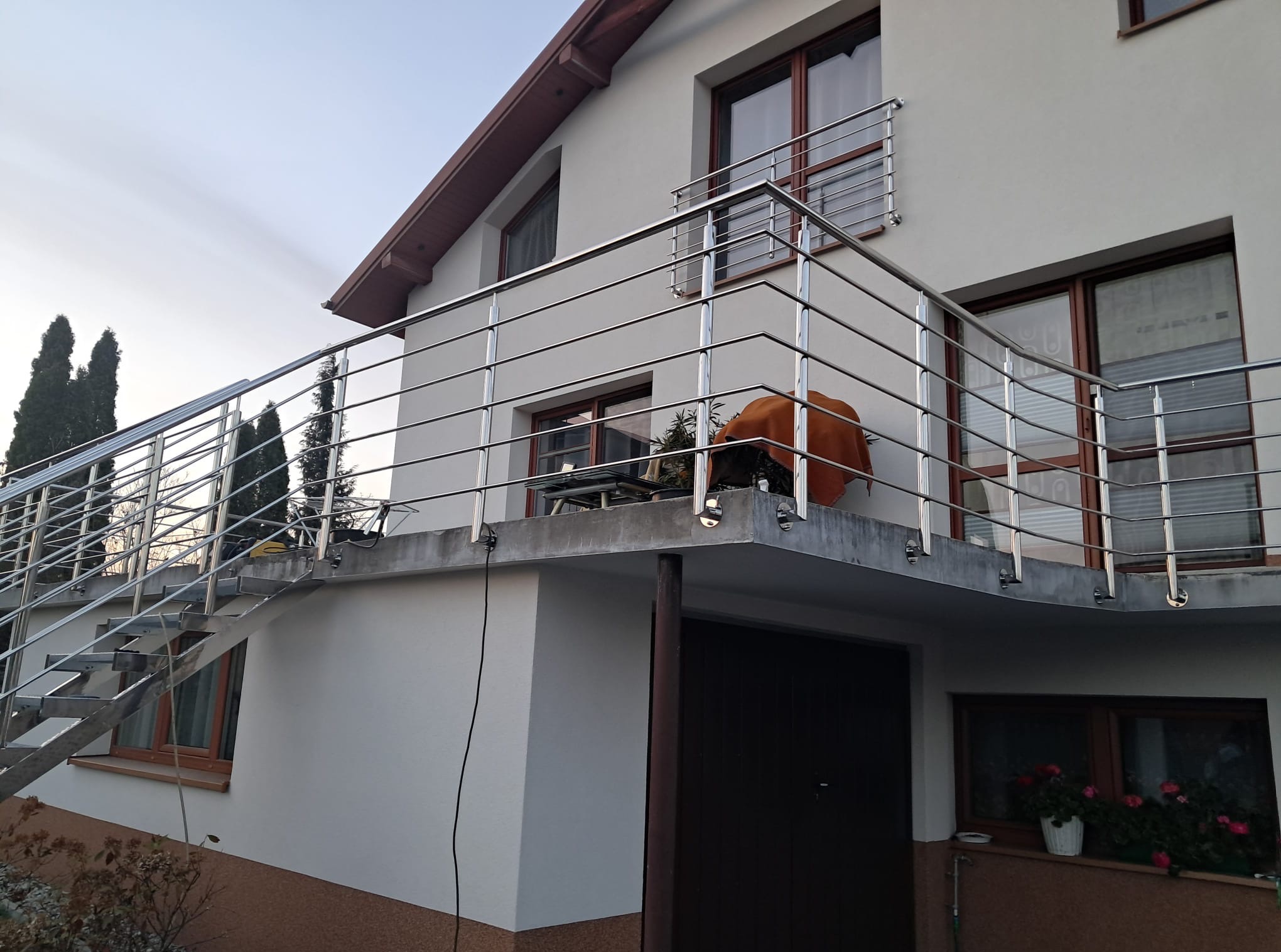 Dom z balkonem i schodami zewnętrznymi, zabezpieczonymi metalową balustradą. Widoczne okna z brązowymi ramami i elementy roślinności. Ujęcie z dołu.