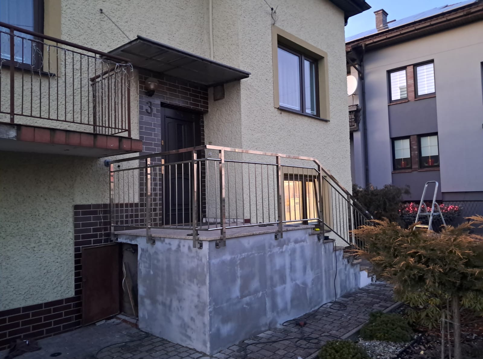 Dom z beżową elewacją i balkonem. Metalowa balustrada w kolorze złota, prowadząca do wejścia. Widoczne schody i ścieżka z kostki brukowej.