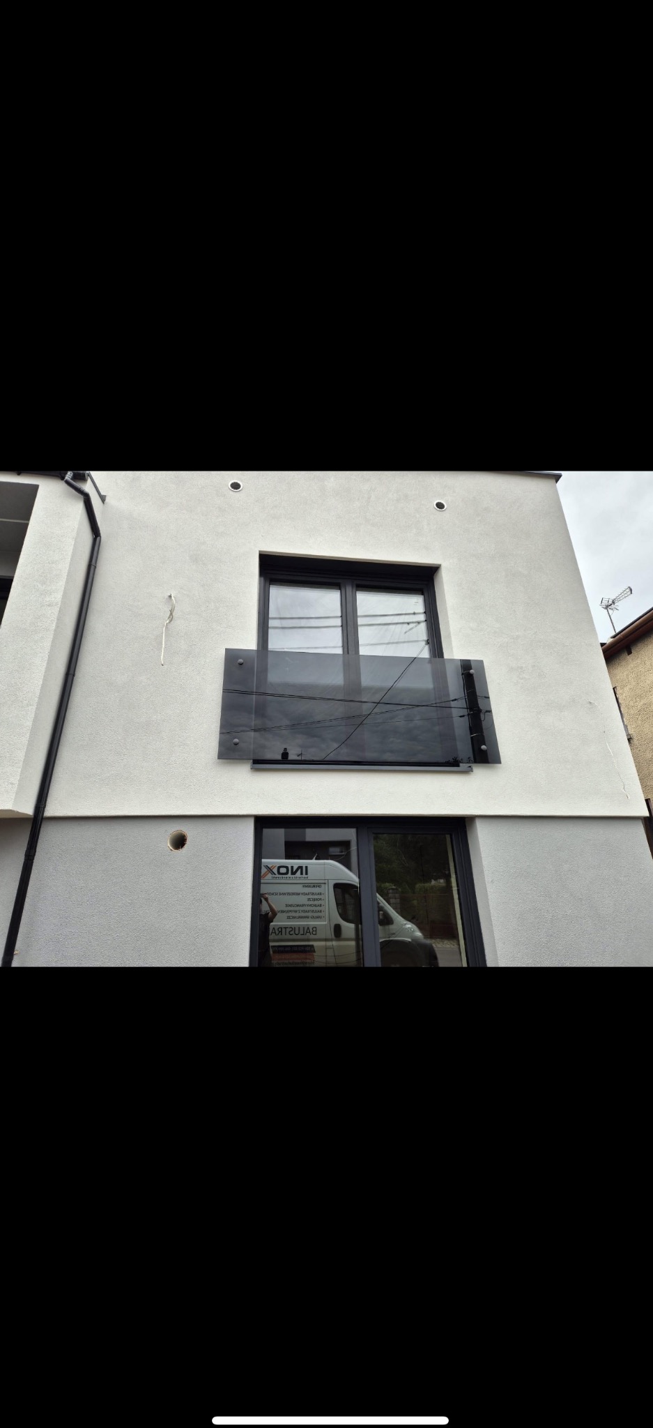 Szklana balustrada balkonowa z ciemnego szkła, zamontowana na nowoczesnym budynku z jasną elewacją. Widoczne mocowania i odbicie otoczenia w szkle.