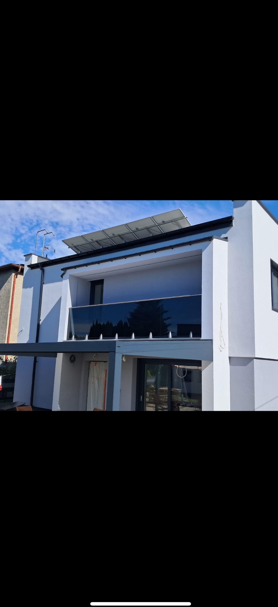 Nowoczesny dom z balkonem z balustradą szklaną, zabezpieczoną kolcami. Na dachu panele słoneczne, zadaszony taras. Biała elewacja, błękitne niebo.