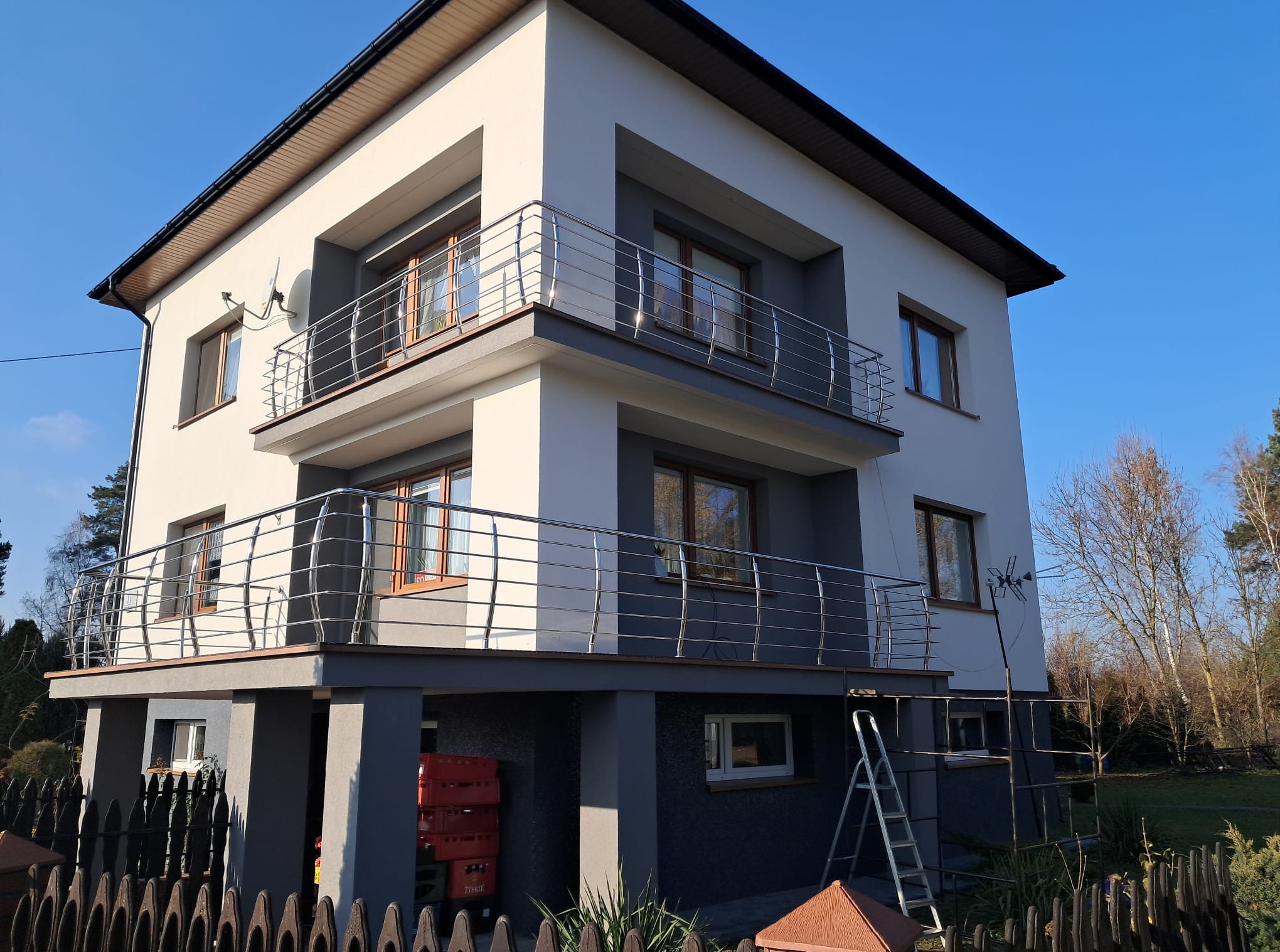 Dwupiętrowy dom z balkonami zabezpieczonymi nowoczesnymi barierkami. Widoczna drabina i skrzynki, co sugeruje prace wykończeniowe. Błękitne niebo w tle.