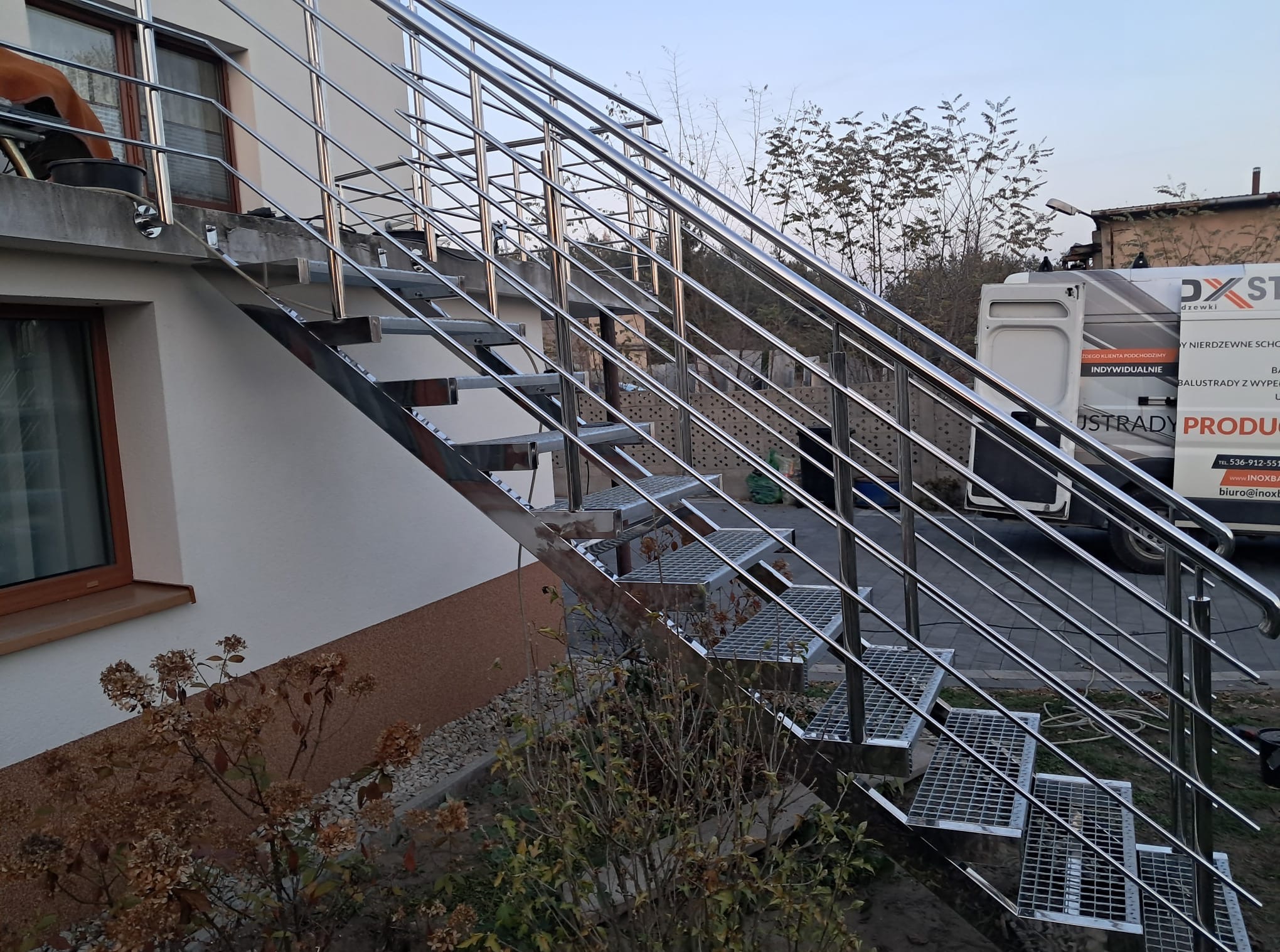 Zewnętrzne schody metalowe z ażurowymi stopniami i poręczami ze stali nierdzewnej, zamontowane przy ścianie budynku. W tle widoczny samochód dostawczy.