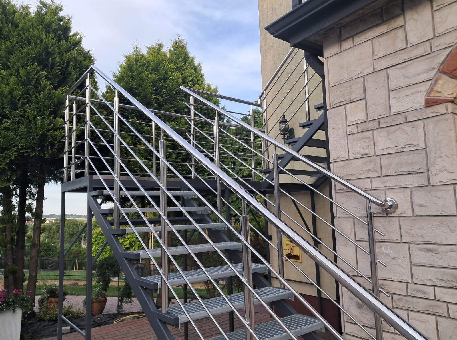 Zewnętrzne schody metalowe z połyskującą balustradą, prowadzące do wejścia budynku z elewacją z kamienia. W tle zieleń drzew i krzewów, błękitne niebo.
