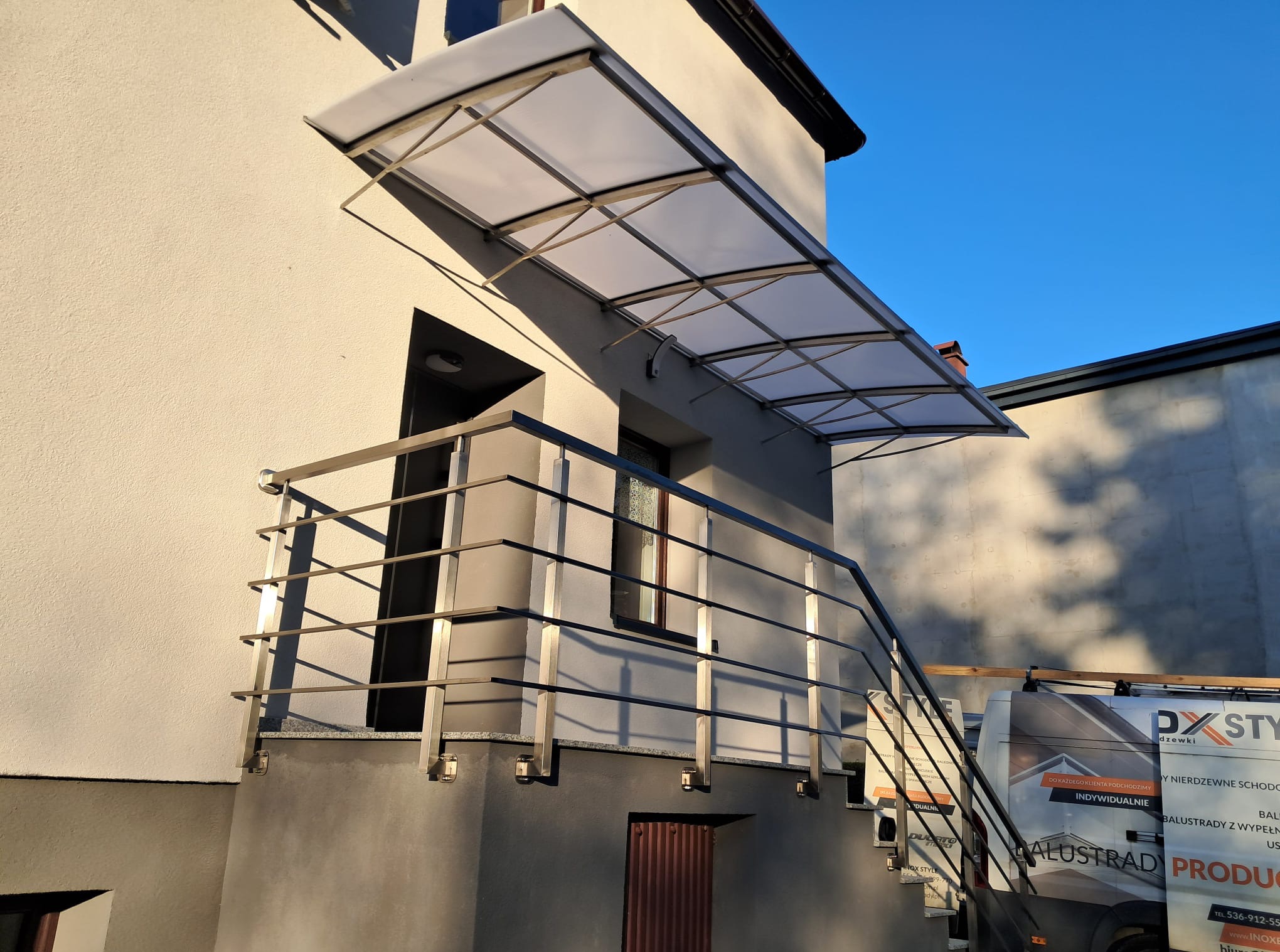 Wejście do budynku z zadaszeniem i metalową balustradą schodową. Widoczny fragment firmowego samochodu dostawczego.