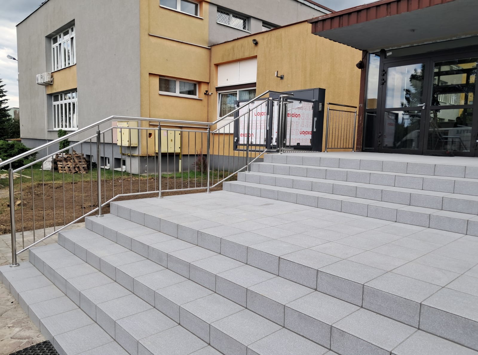 Zewnętrzne schody z szarych płytek z metalową poręczą i podjazdem dla niepełnosprawnych na tle budynku w odcieniach szarości i żółci.