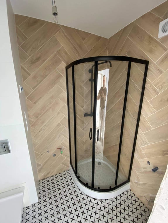 Wykończona łazienka z kabiną prysznicową w czarnej ramie, beżowymi płytkami ułożonymi w jodełkę na ścianach i wzorzystą podłogą. Widoczne elementy armatury i wentylacja.