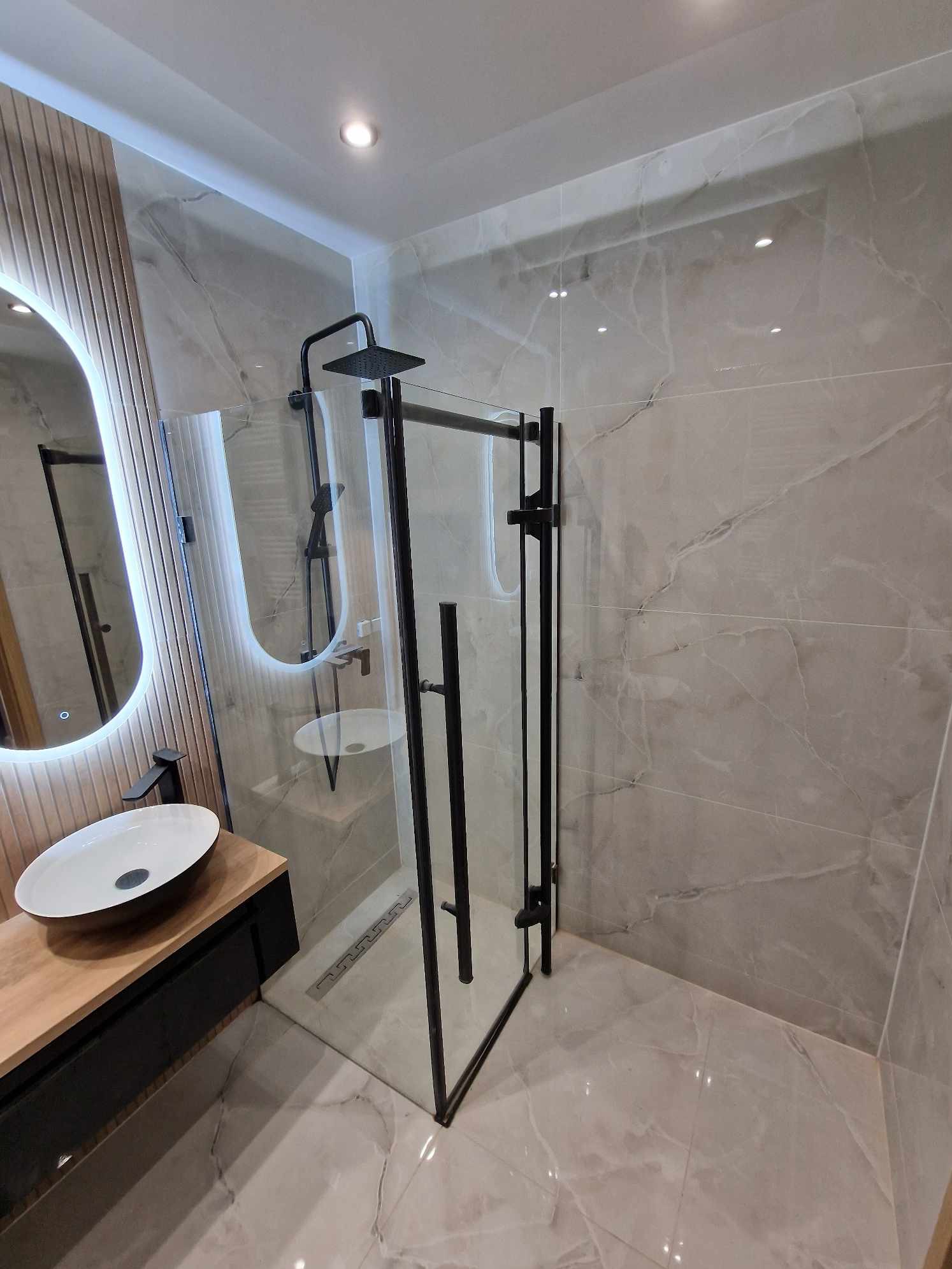 Nowoczesna łazienka z kabiną prysznicową typu walk-in z czarnymi profilami, umywalką nablatową i podświetlanym lustrem. Marmurowe płytki na ścianach i podłodze.